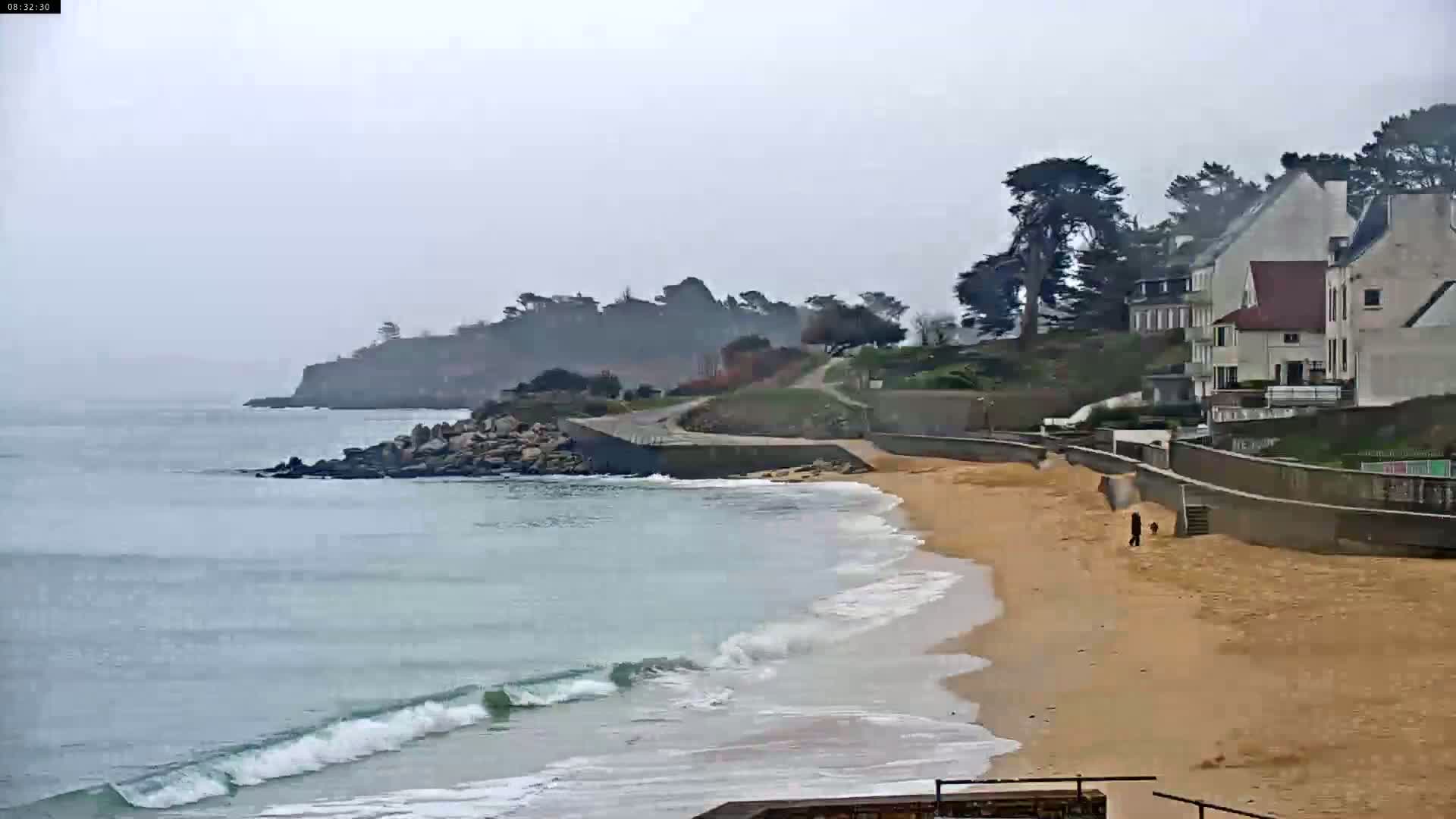 A sandy beach with gentle waves laps the shore under an overcast sky, near a rocky cove and a line of houses on a hillside.