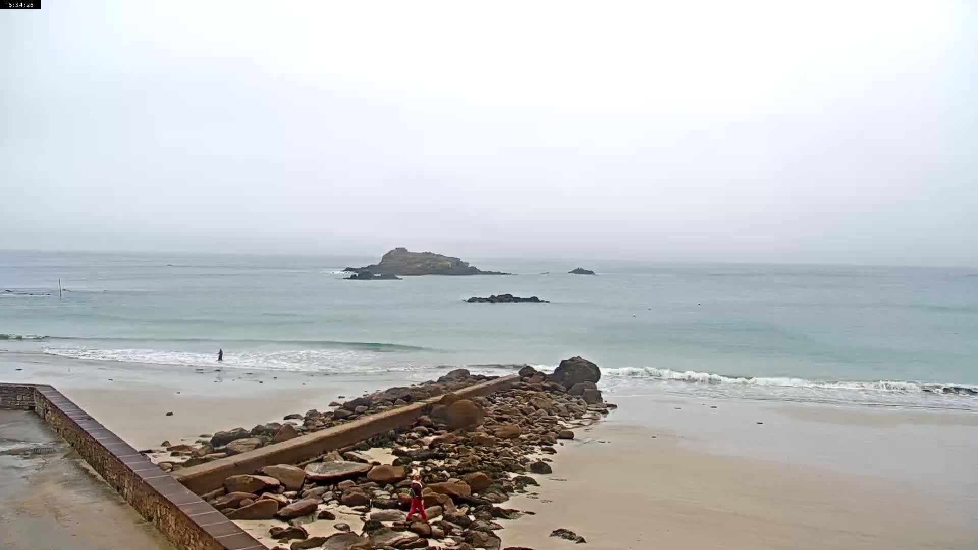 A sandy beach with a rocky jetty extends into a calm, light-green ocean under an overcast sky, with several small islands visible in the distance.
