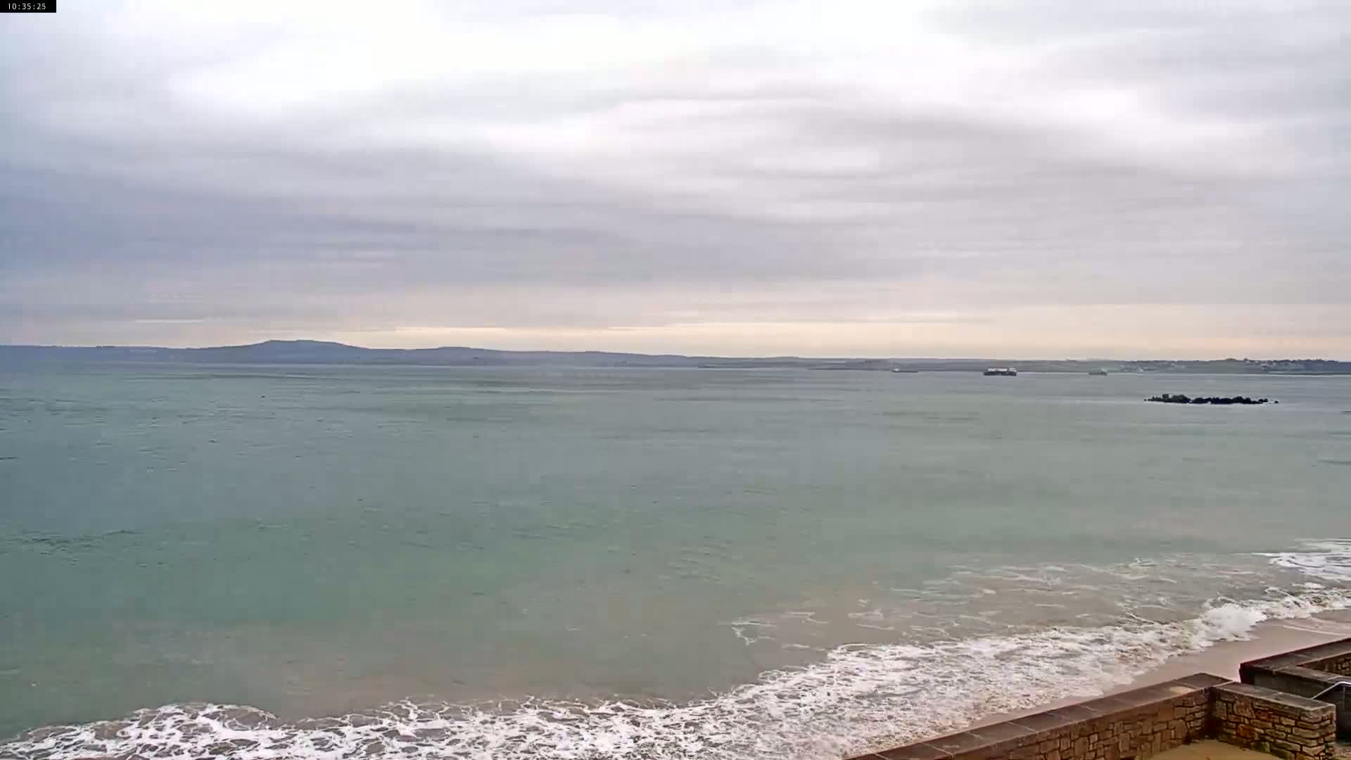 A calm sea with small waves on a beach under a cloudy sky, with a distant landmass visible on the horizon.