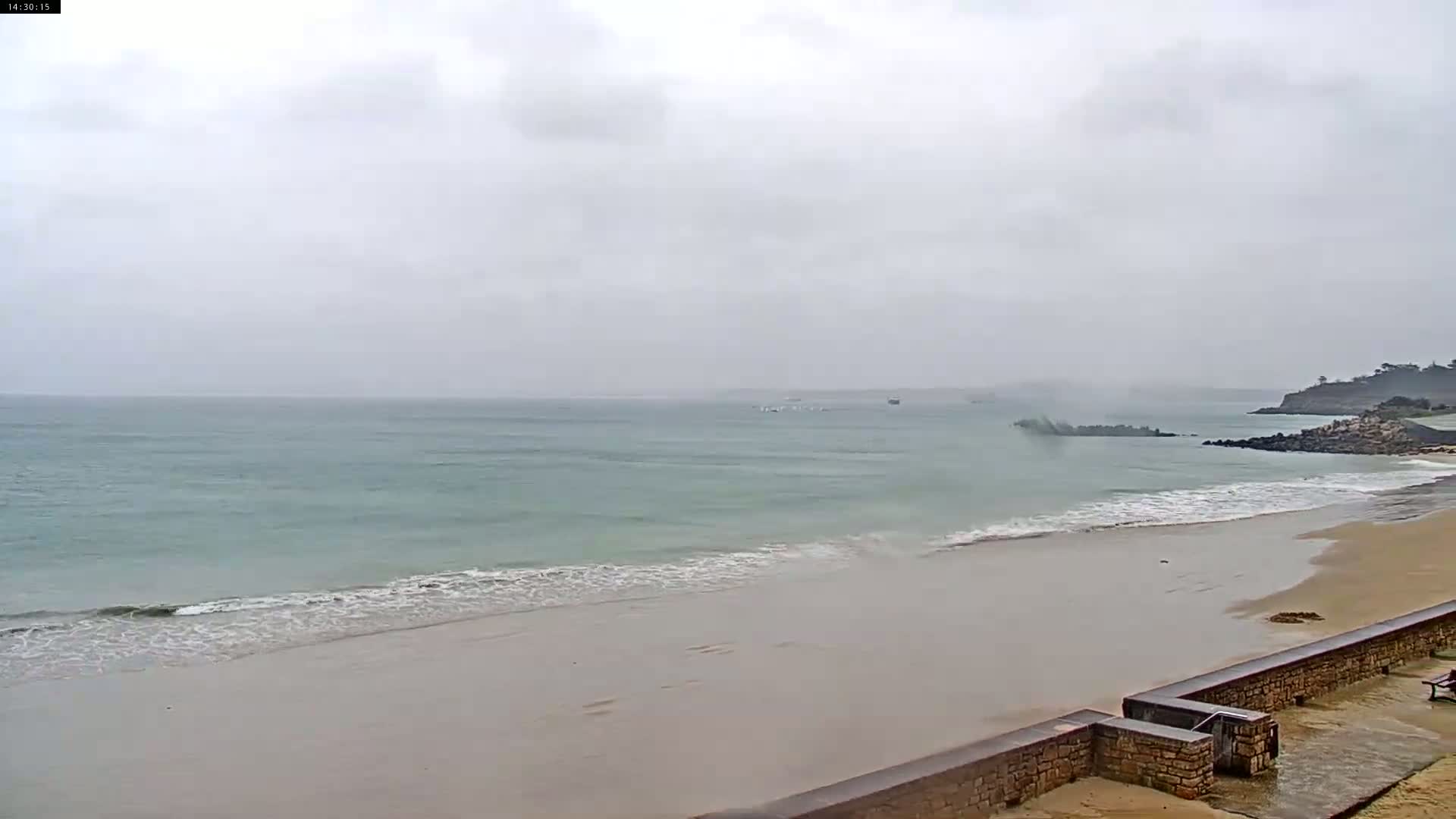 A mostly overcast sky hangs over a calm ocean meeting a sandy beach, partially bordered by a low stone wall.