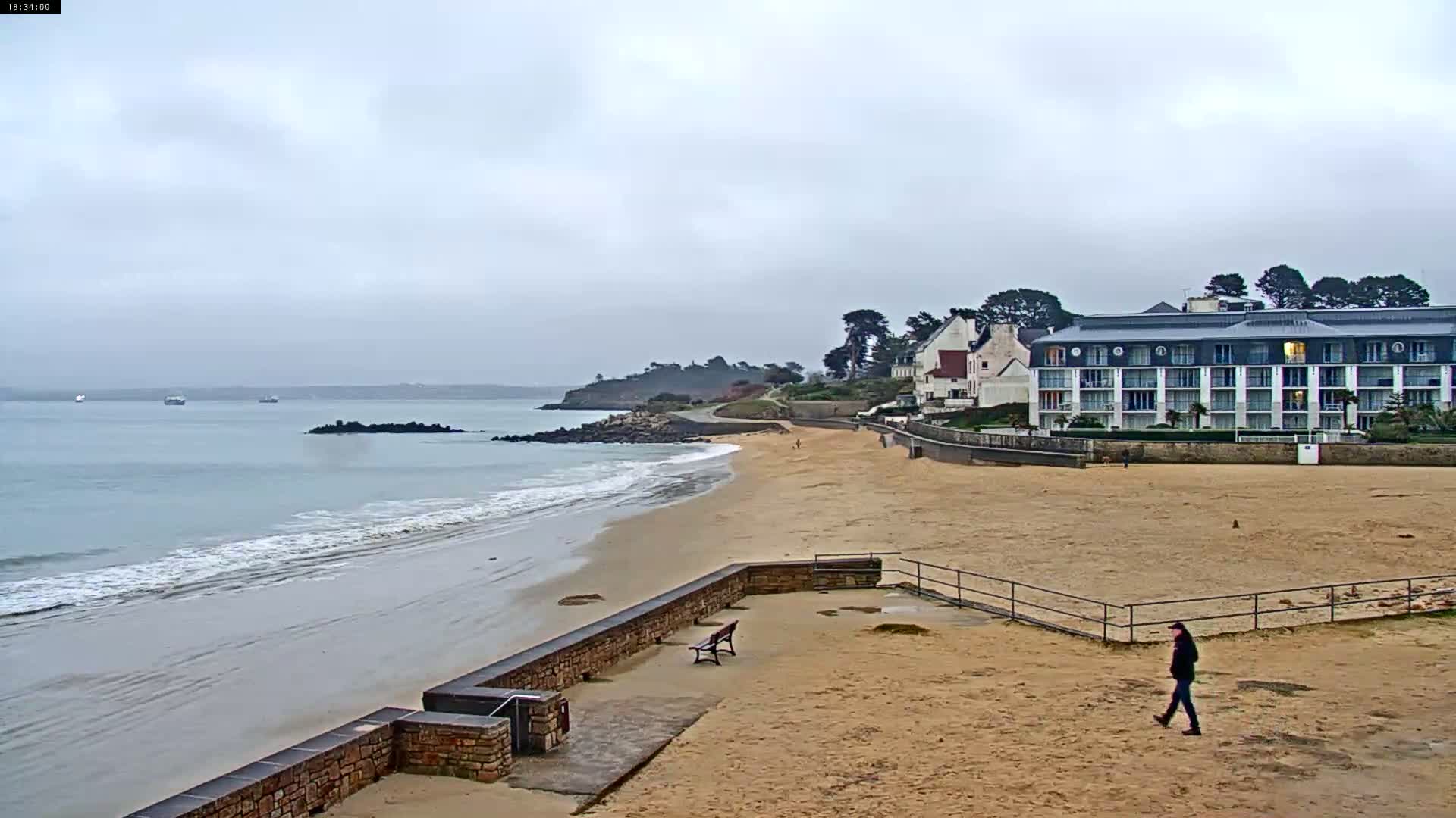 A sandy beach with gentle waves under an overcast sky, features a stone wall, a bench, a few people, and a large building in the background.