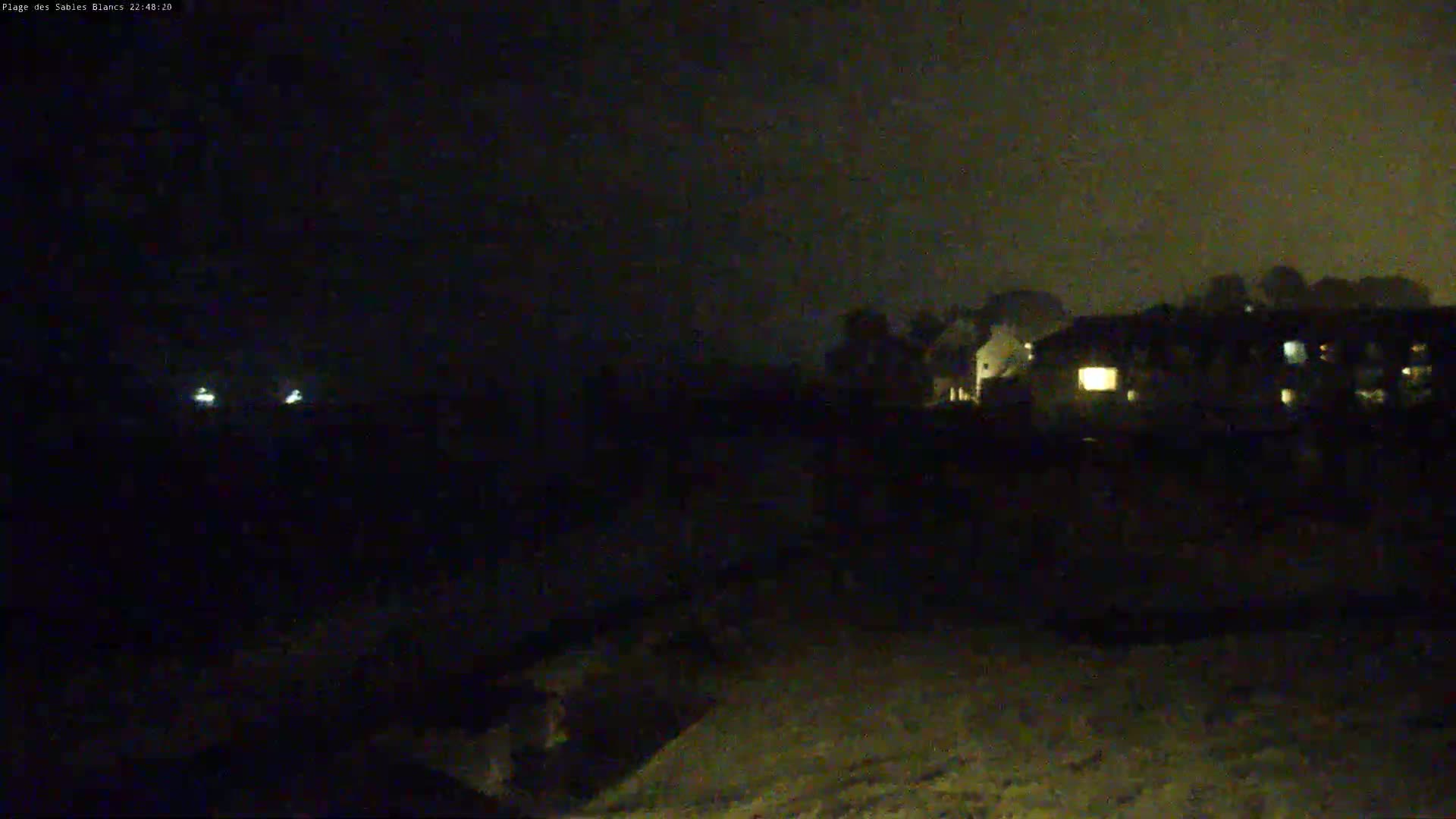 A dark, nighttime scene shows a line of houses with a few lights on, situated beyond a mostly dark foreground under a cloudy sky.