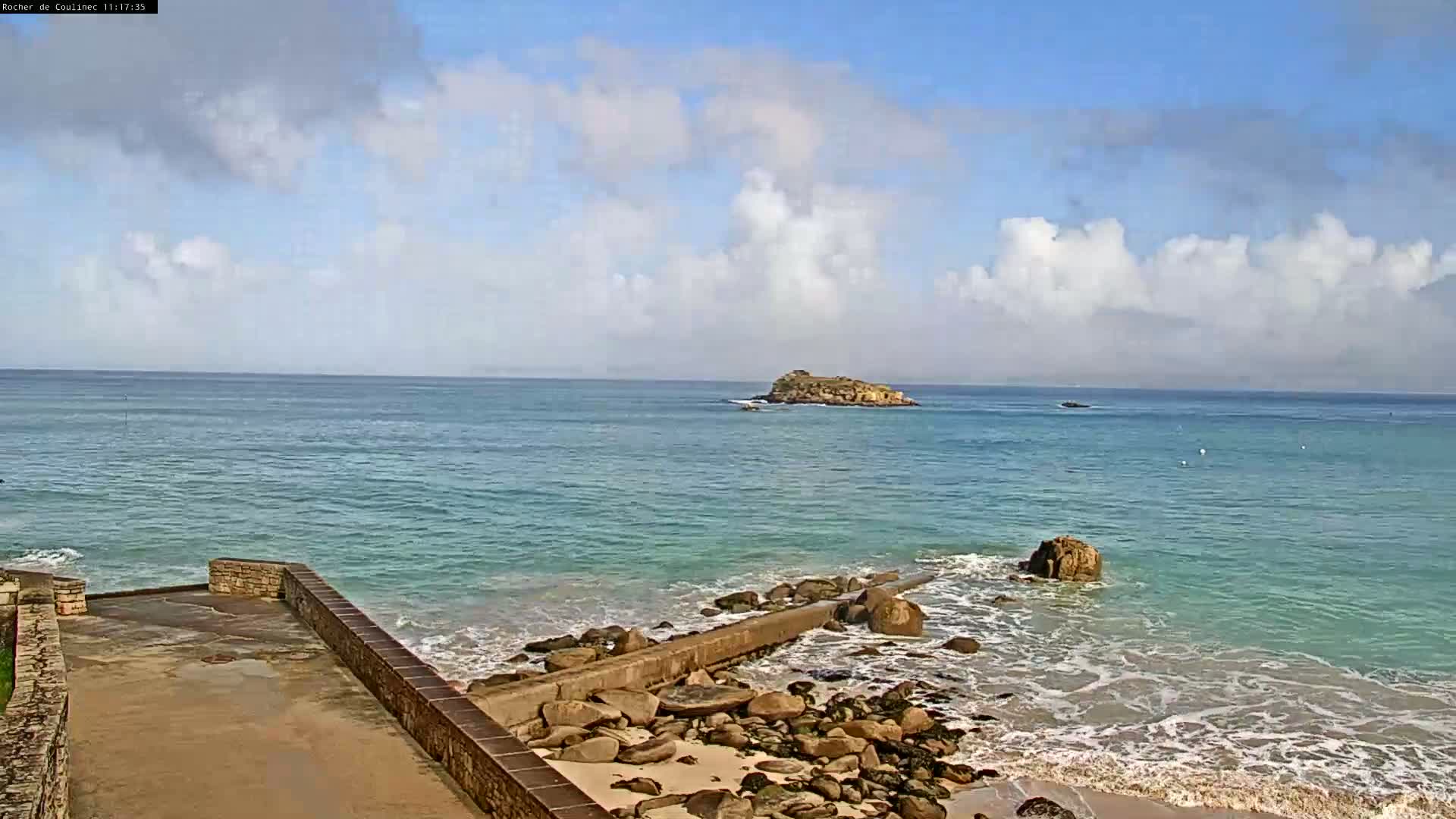 A rocky shoreline with a low stone wall leads to a calm, teal ocean with a small island visible in the distance under a partly cloudy sky.