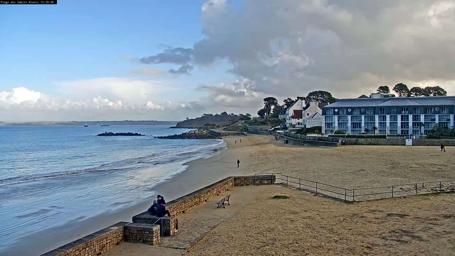 A sandy beach with calm waves, a stone wall, a few people, and a multi-story building is visible under a partly cloudy sky.