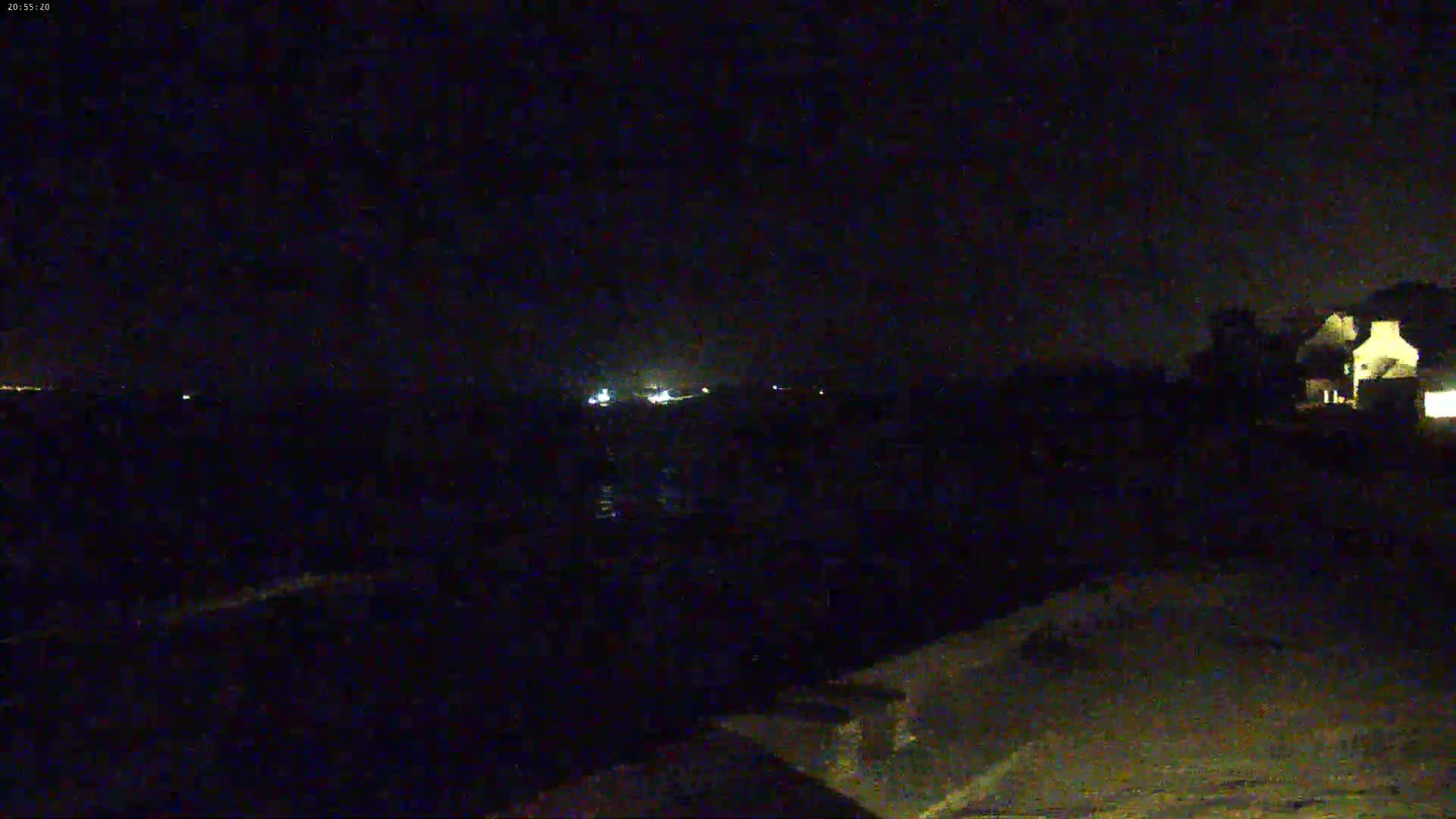 A nighttime, low-light view shows a dark body of water, a dimly lit shoreline, and a building with illuminated windows in the upper right corner.