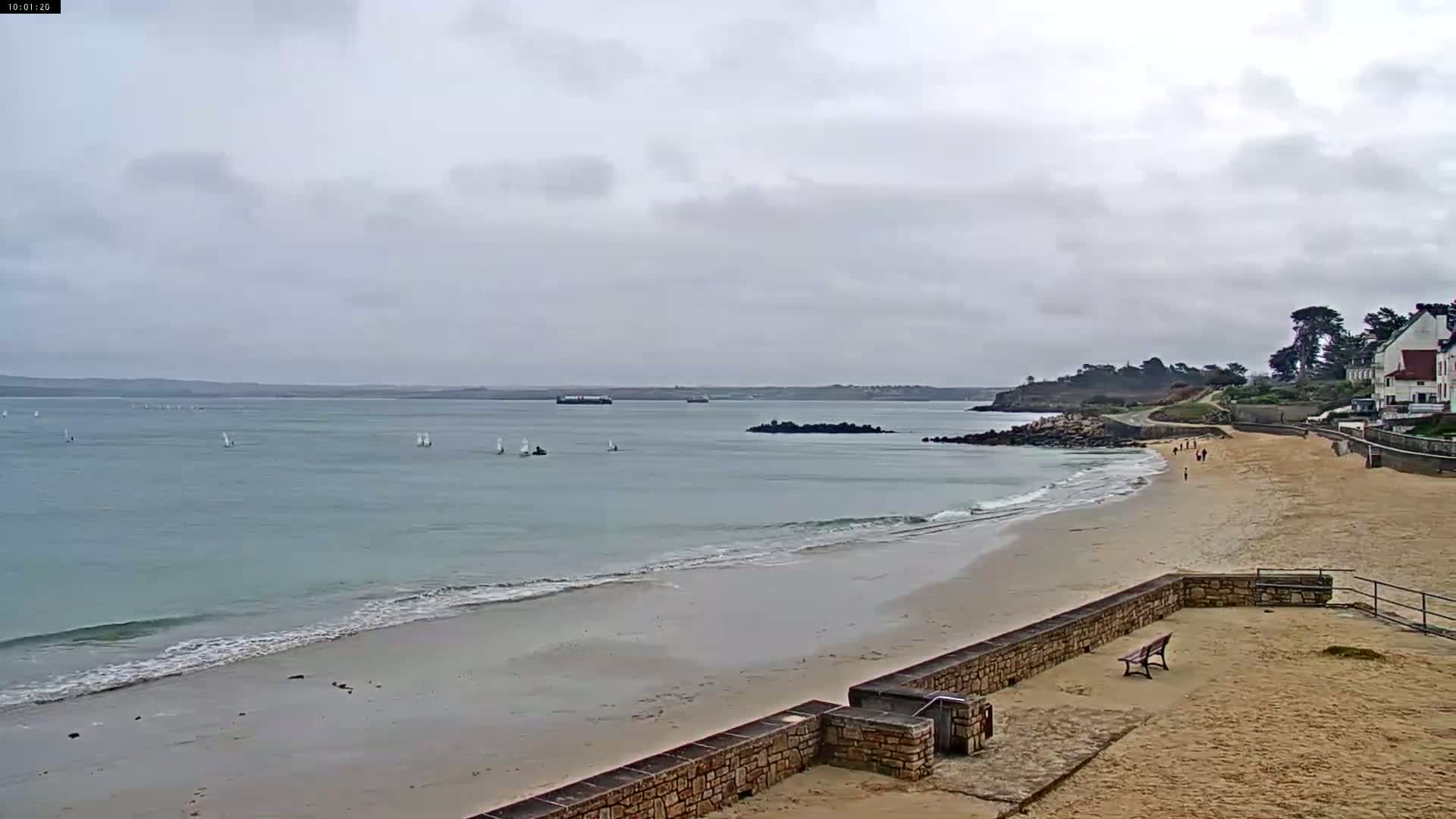 A sandy beach with a stone wall and a few people stretches to a calm ocean with several sailboats under a cloudy sky.