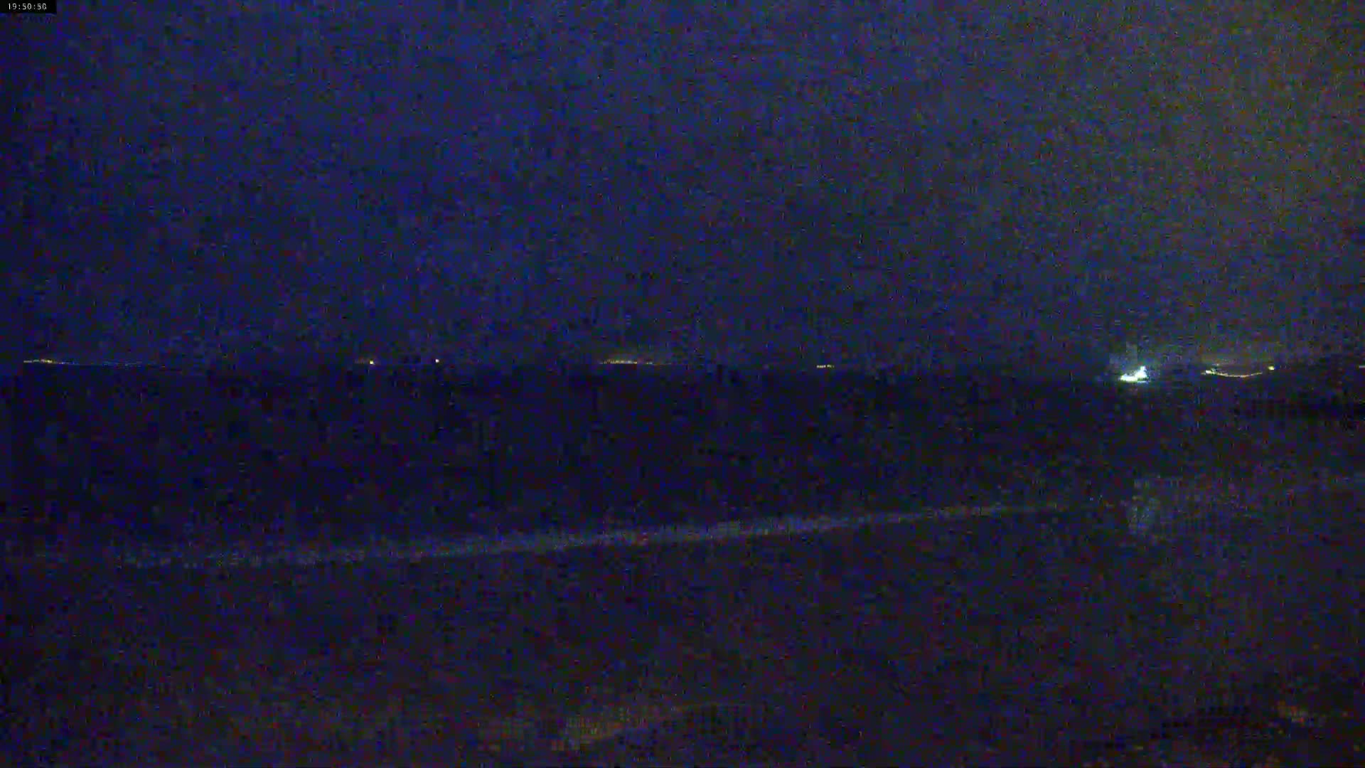 A dark, nighttime scene shows a distant shoreline with faint lights under a dark, cloudy sky.