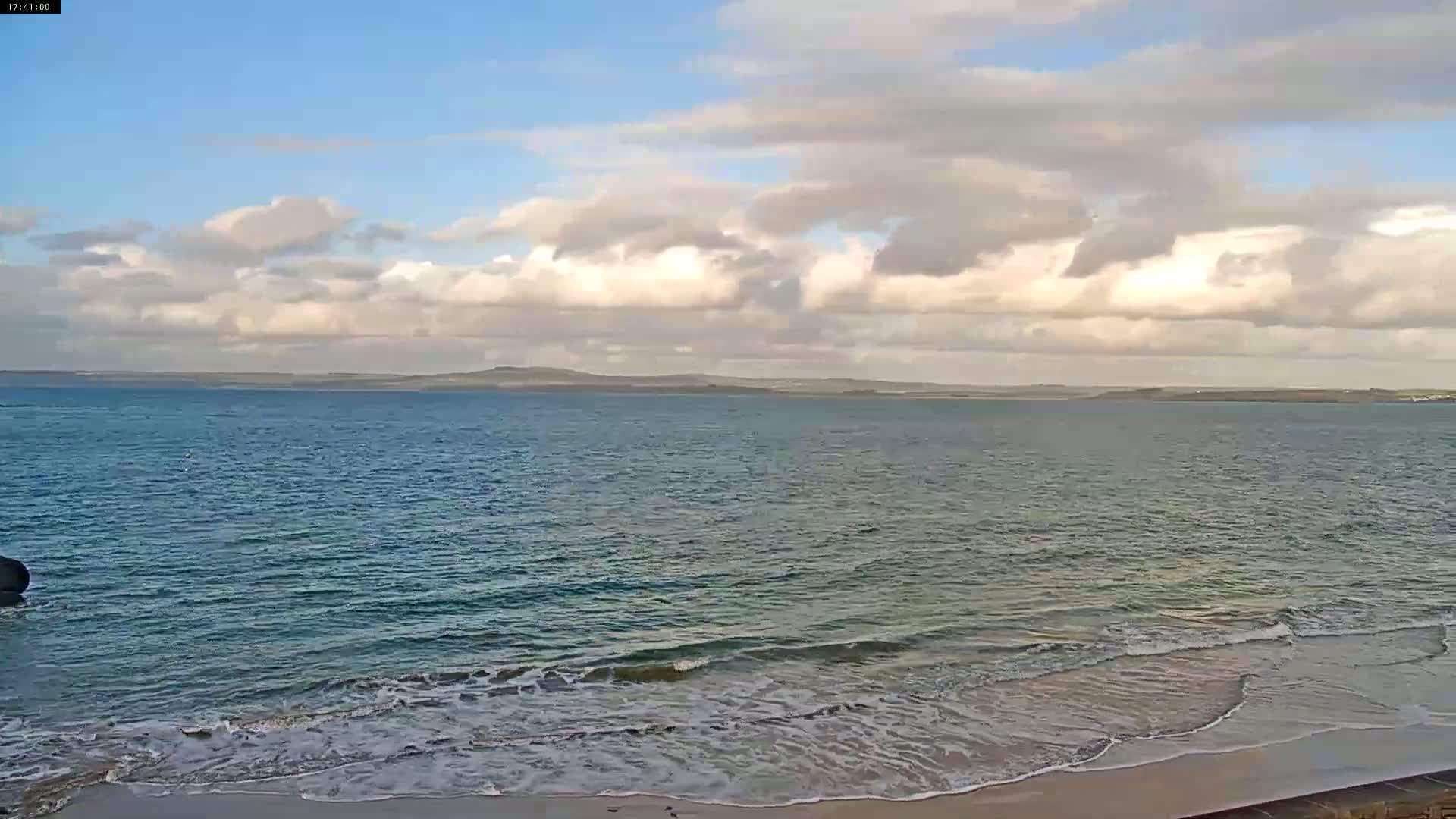 A partly cloudy day shows a calm ocean with small waves lapping a sandy beach, and a distant landmass visible on the horizon.