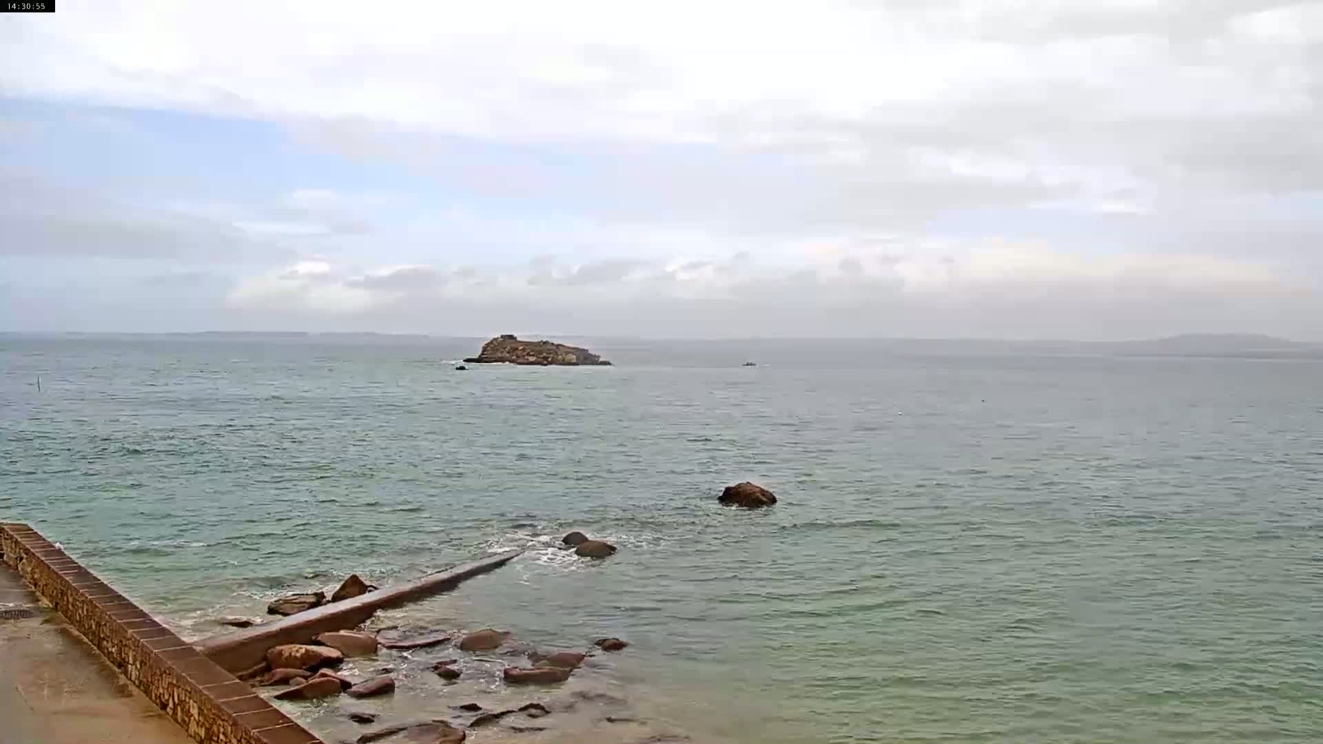 A rocky shoreline with a low wall extends into a calm, somewhat overcast ocean where a small island is visible in the distance.