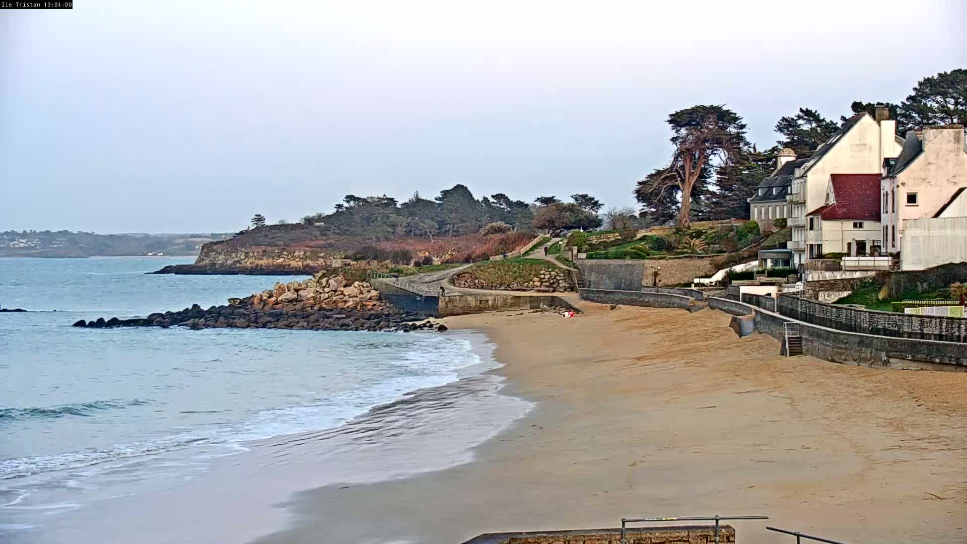 A sandy beach with gentle waves laps the shore near a rocky coastline, where several houses sit on a hill overlooking the water under a cloudy sky.