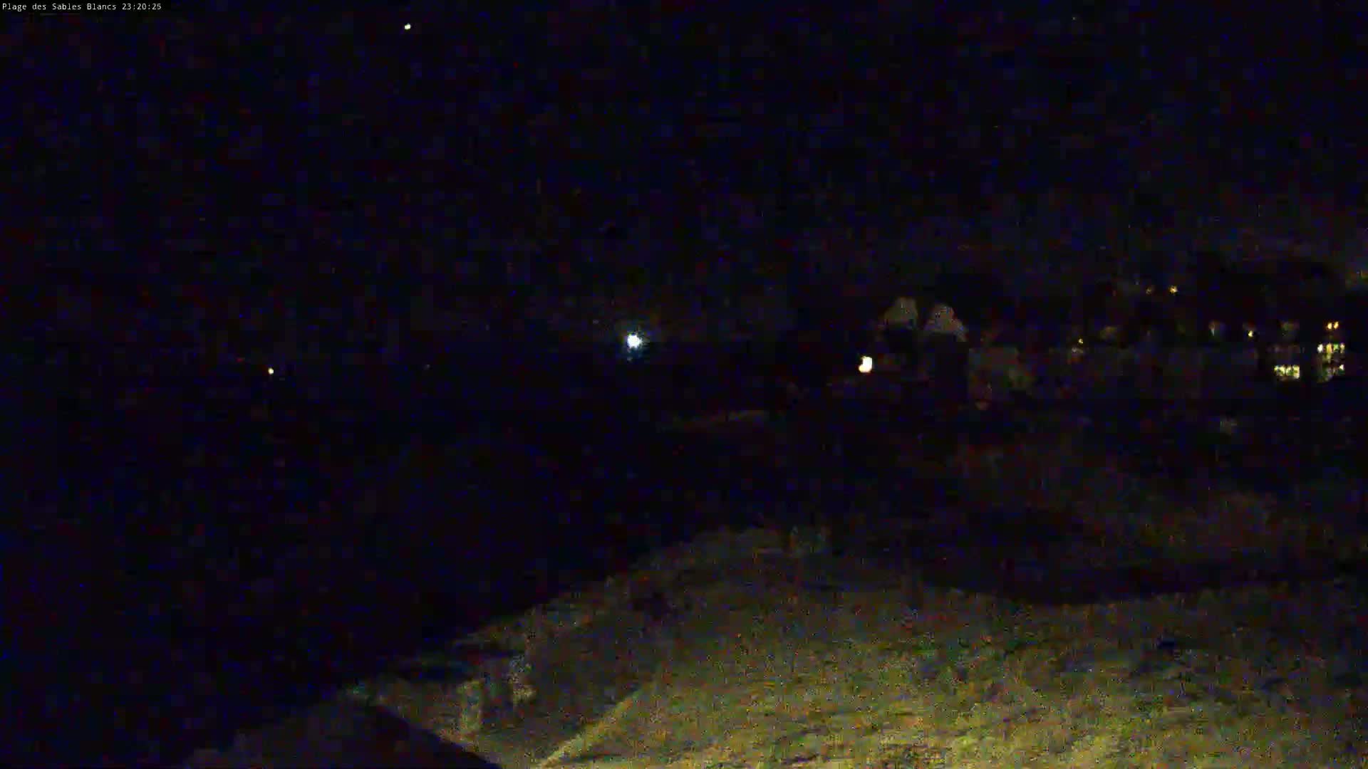 A nighttime, low-light view of a dark landscape with a few faint lights in the distance, possibly buildings, under a mostly clear, starlit sky.