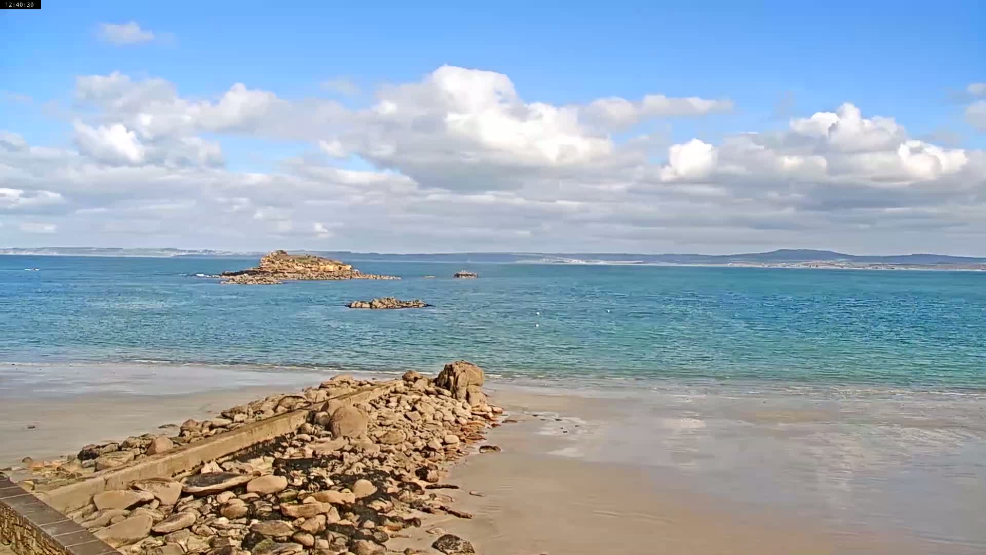 A sandy beach meets a rocky outcrop leading to a calm, turquoise ocean under a partly cloudy sky, with islands visible in the distance.