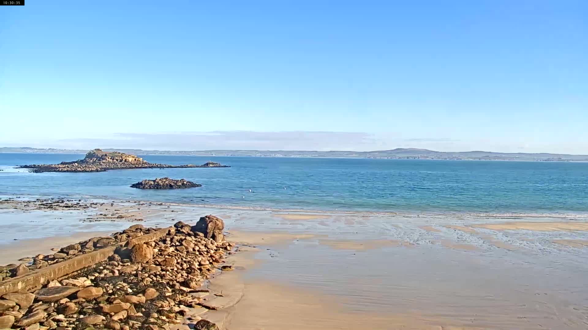 A sandy beach with rocky outcrops and a calm, blue ocean stretches to a distant landmass under a clear blue sky.