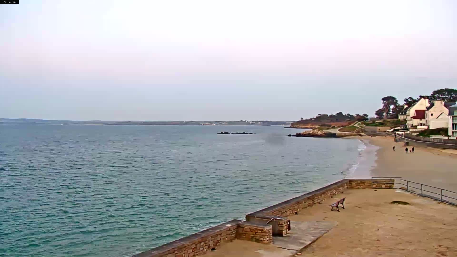 A calm, light-blue sea laps a sandy beach, where a few people stroll, near a low stone wall and some houses on a cloudy day.