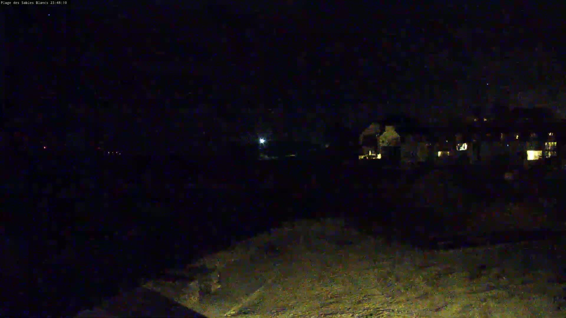 A nighttime, low-light view shows a dark, mostly empty landscape with some dimly lit buildings in the distance and a few faint lights in the foreground.