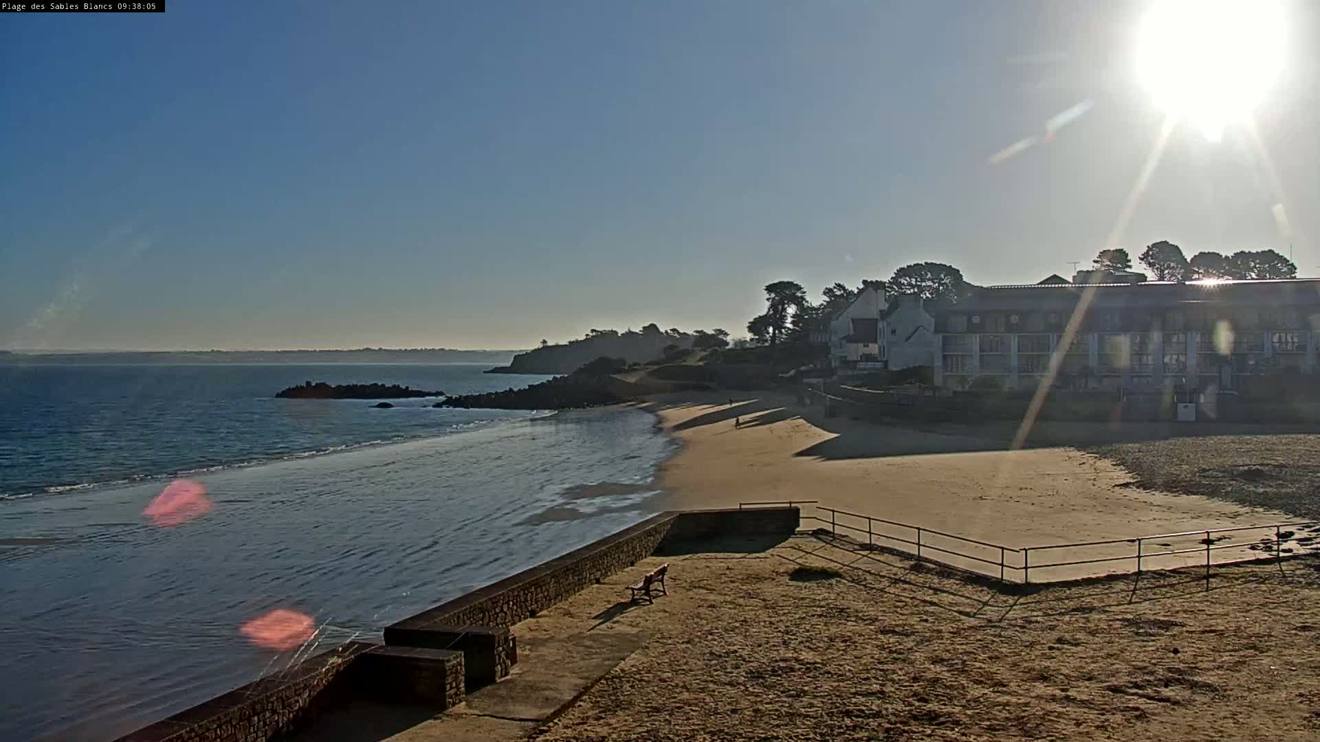 A sandy beach with calm, shallow water stretches out to a calm ocean under a bright, sunny sky, with a building and some trees visible in the background.