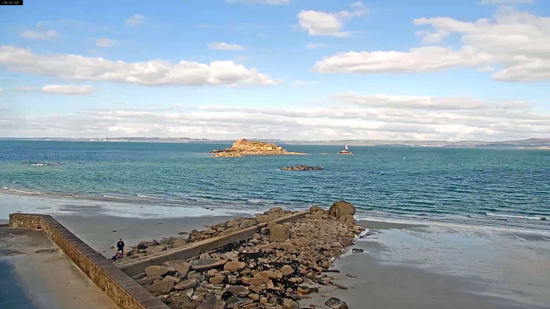 A sandy beach with a rocky jetty stretches toward a calm, blue ocean under a partly cloudy sky, featuring a small island in the distance.