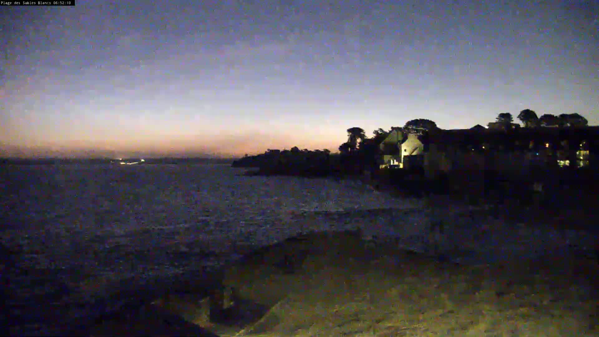A dark, slightly wavy ocean meets a shoreline with silhouetted buildings under a twilight sky.