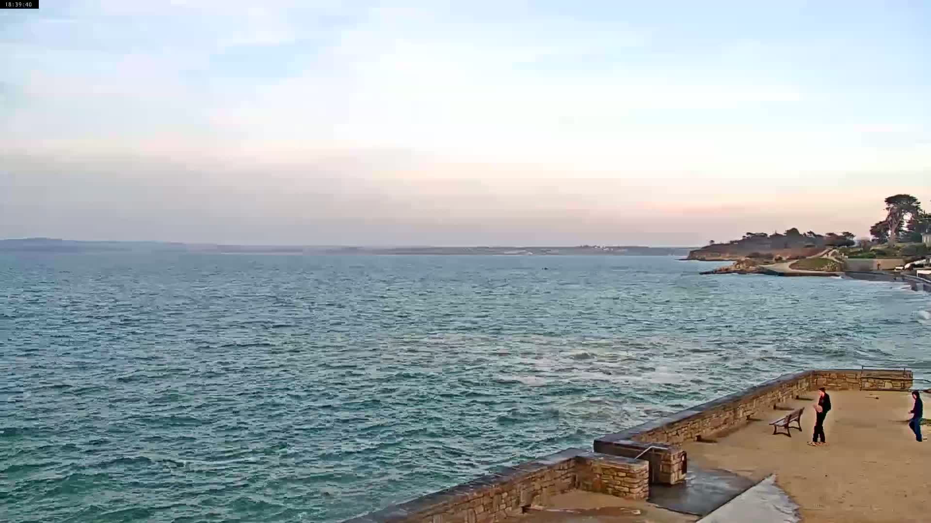 Two people stand on a stone wall overlooking a choppy ocean under a mostly clear sky.