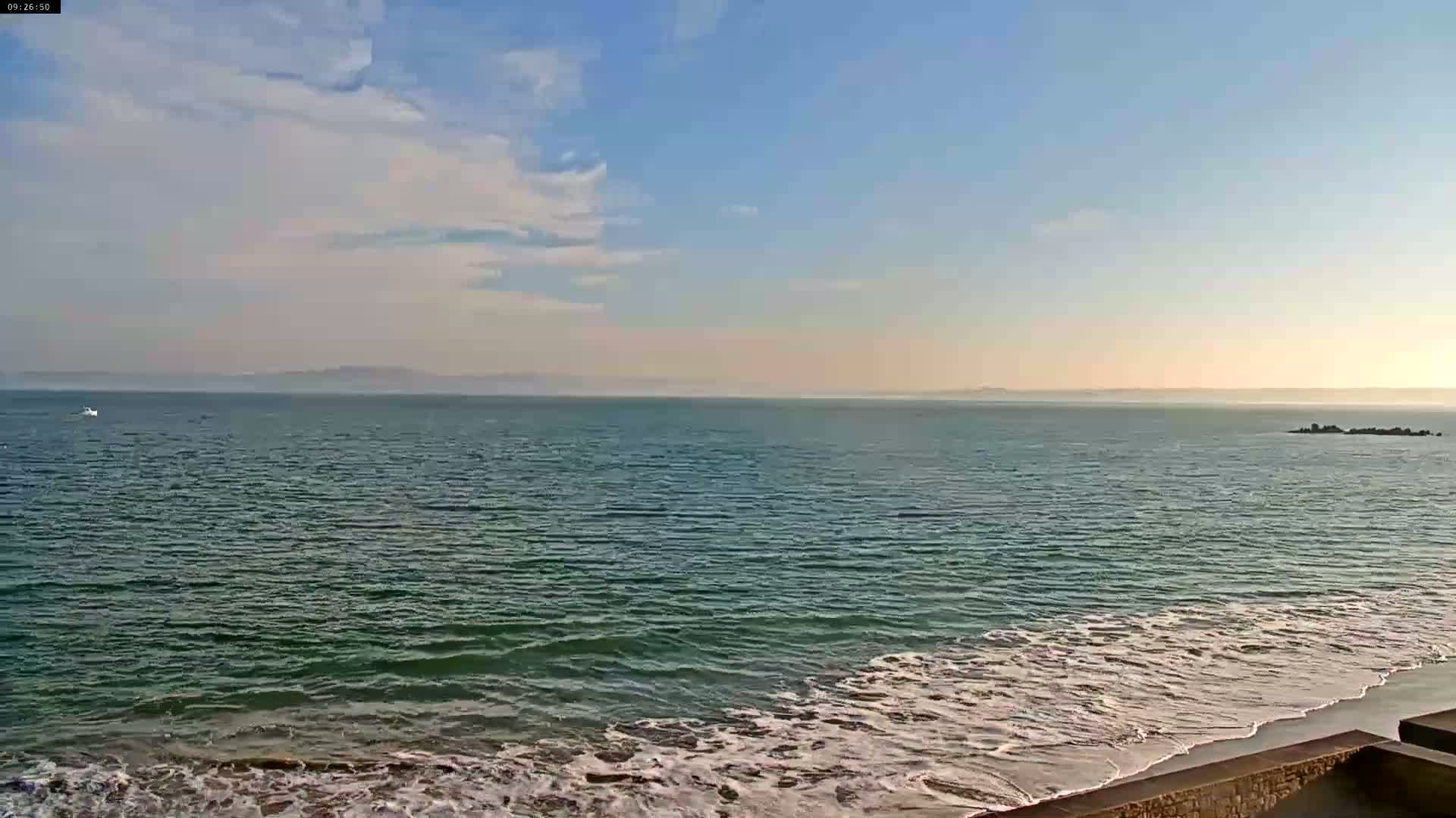 A mostly sunny day shows a calm ocean with small waves lapping a sandy beach, with a distant coastline visible on the horizon.
