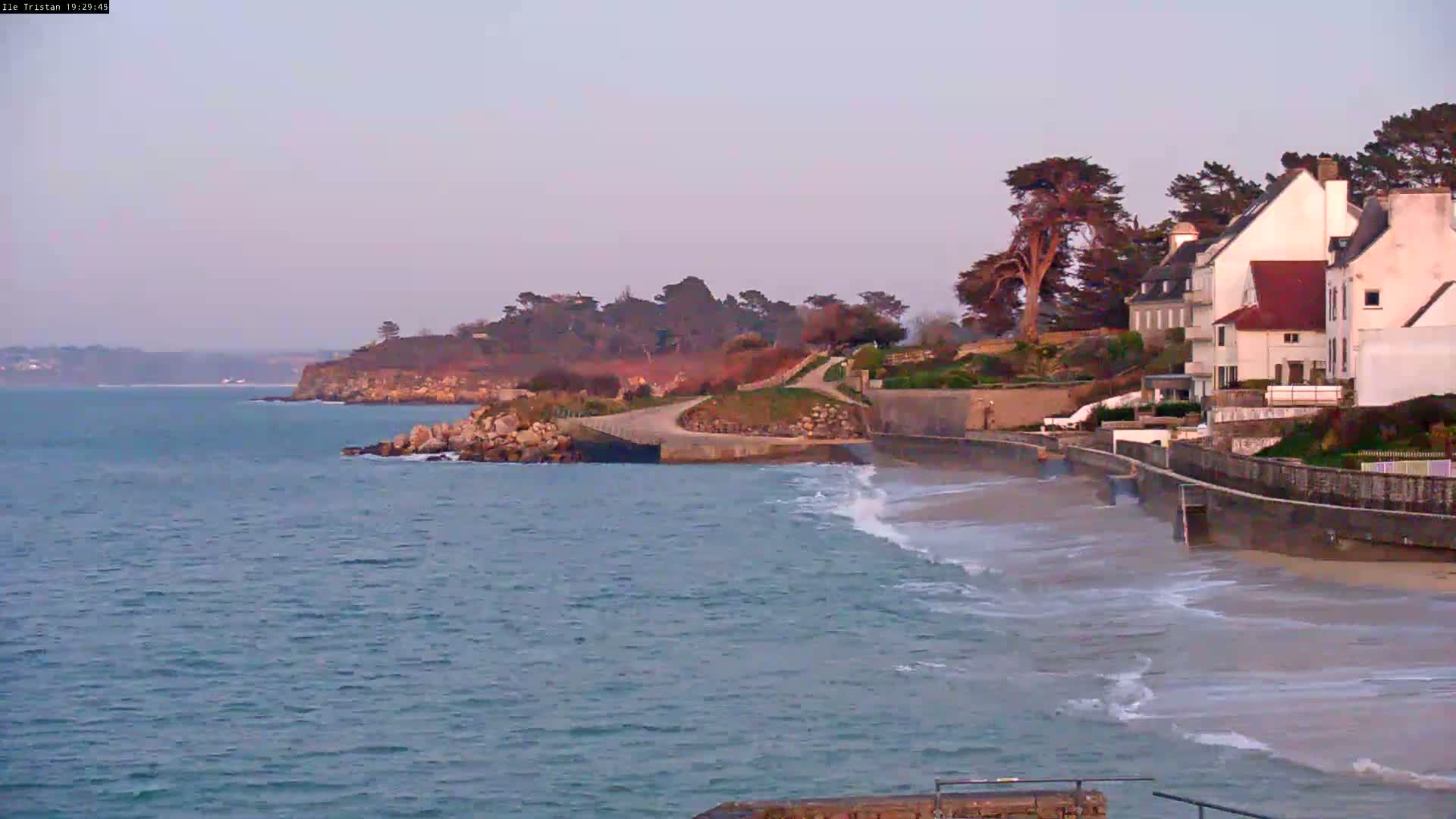 A coastal scene at dusk shows a calm sea lapping a sandy beach beside a seawall, behind which several houses are situated on a rocky headland.