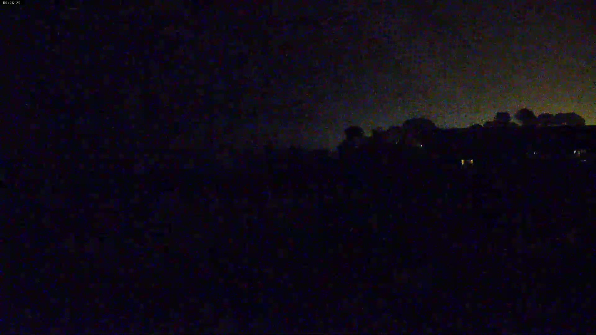 A dark, nighttime scene shows a distant line of trees and buildings under a slightly cloudy sky.