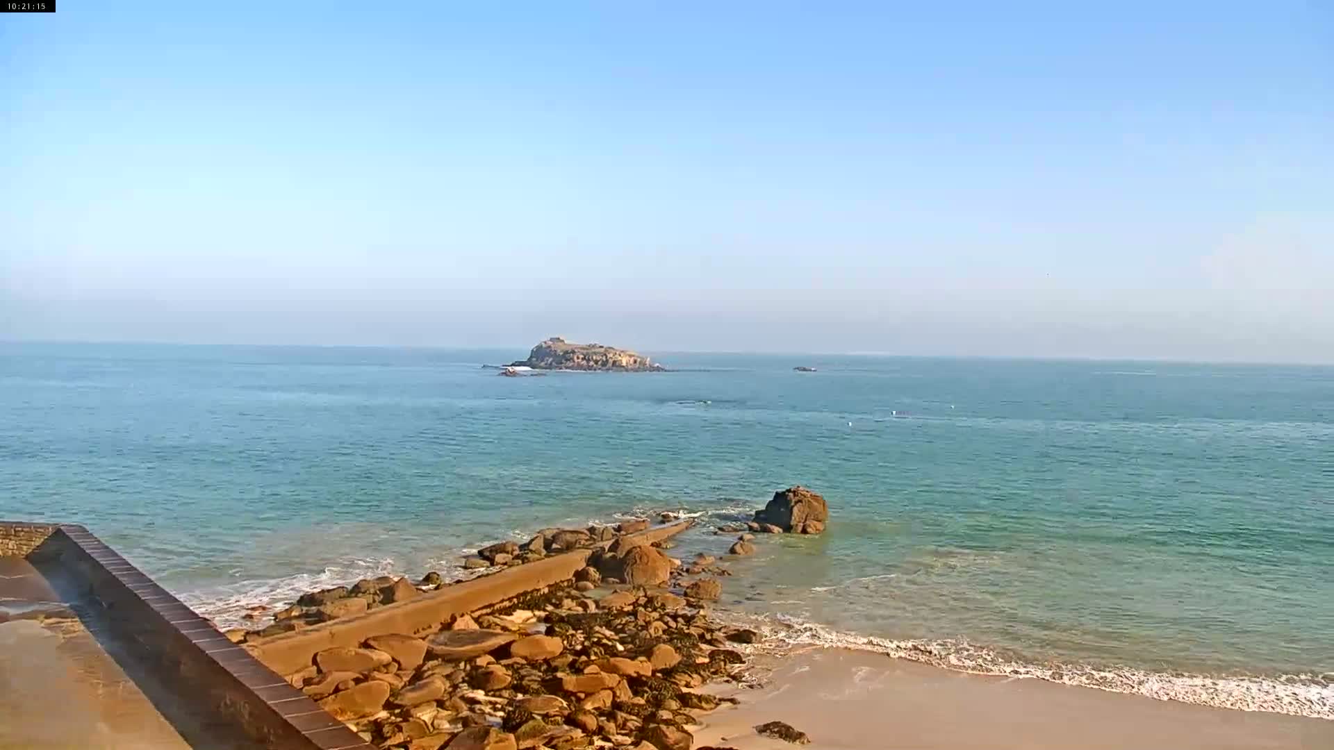 A rocky shoreline with a low wall extends into a calm, clear ocean toward a small island under a clear blue sky.