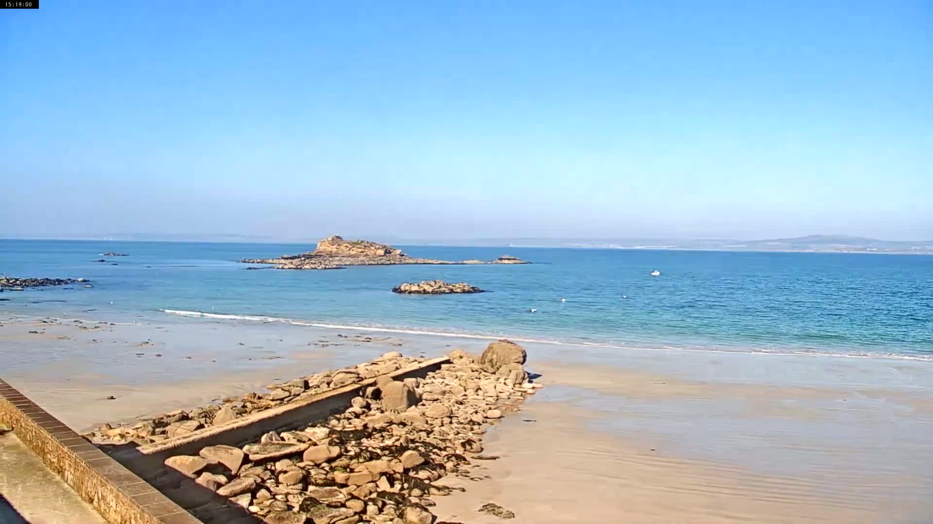 A sandy beach with calm, light blue water meets a rocky shoreline extending to a small island under a clear blue sky.