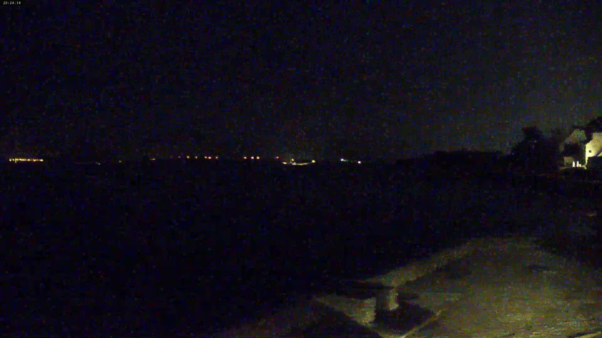 A nighttime, outdoor scene shows a dark landscape with a line of distant lights and a partially visible building to the right.