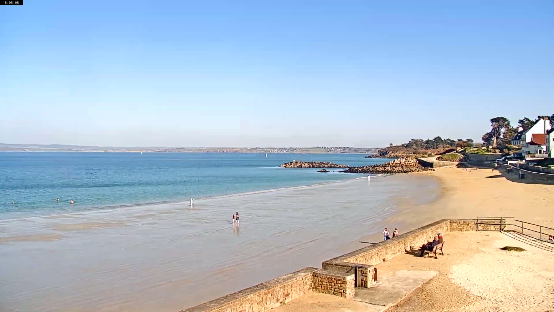 A sandy beach stretches out to a calm, light blue ocean under a clear blue sky, with a few people scattered on the sand and in the water near a rocky outcrop and some buildings.