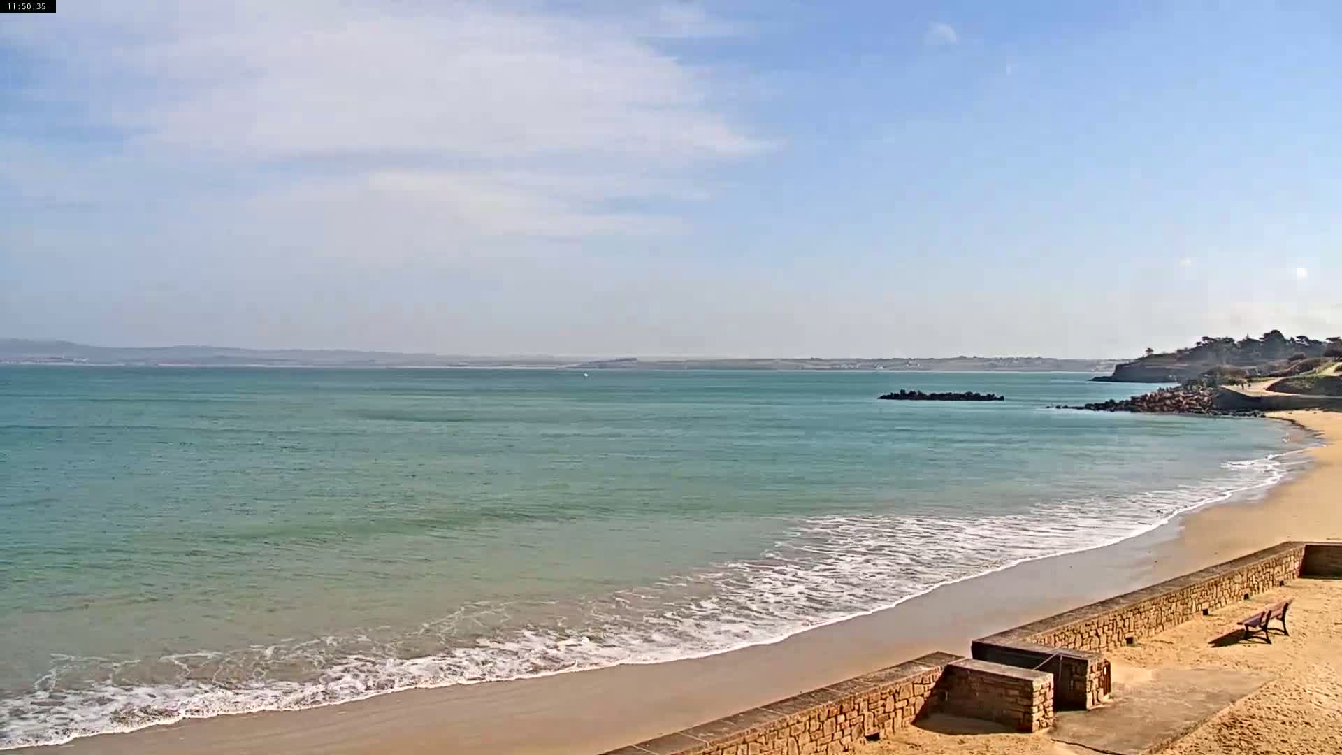 A sandy beach with gentle waves laps the shore under a mostly sunny sky, with a low-lying landmass visible in the distance.
