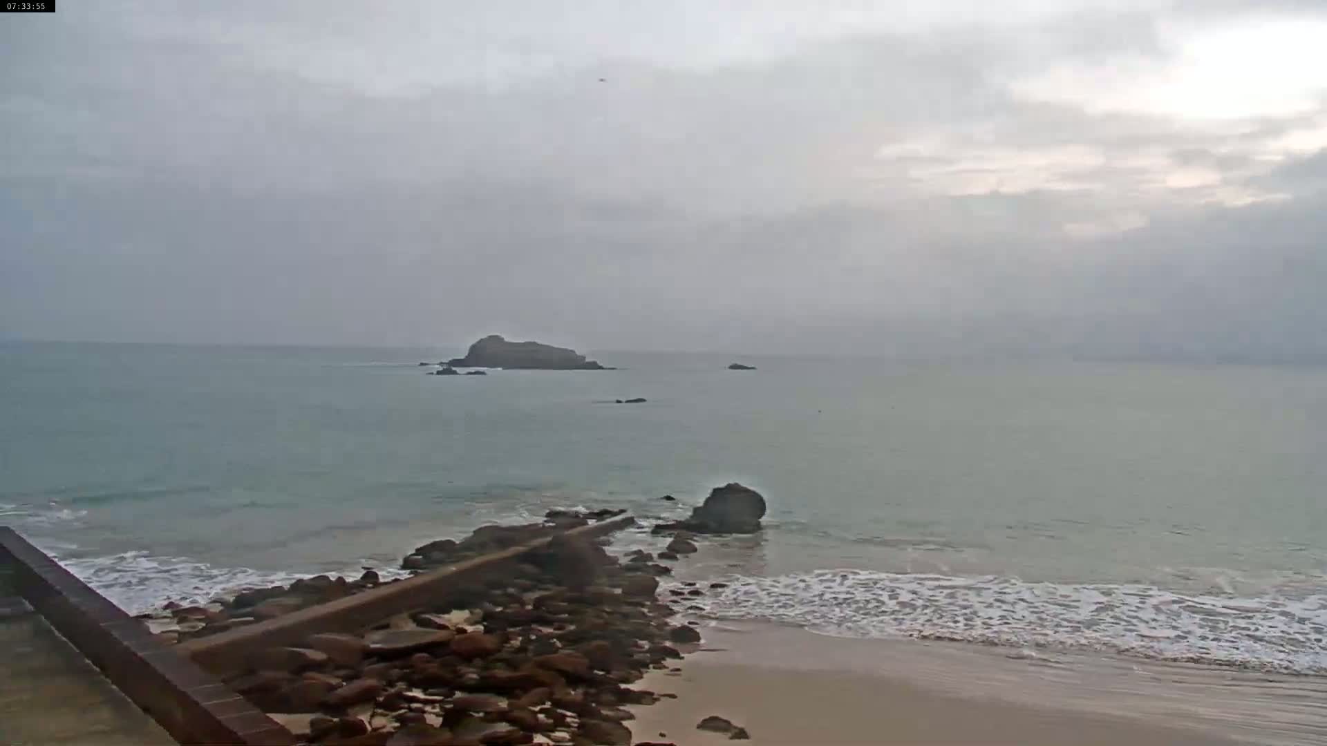 A rocky shoreline meets a calm, grey sea under an overcast sky, with small islands visible in the distance.