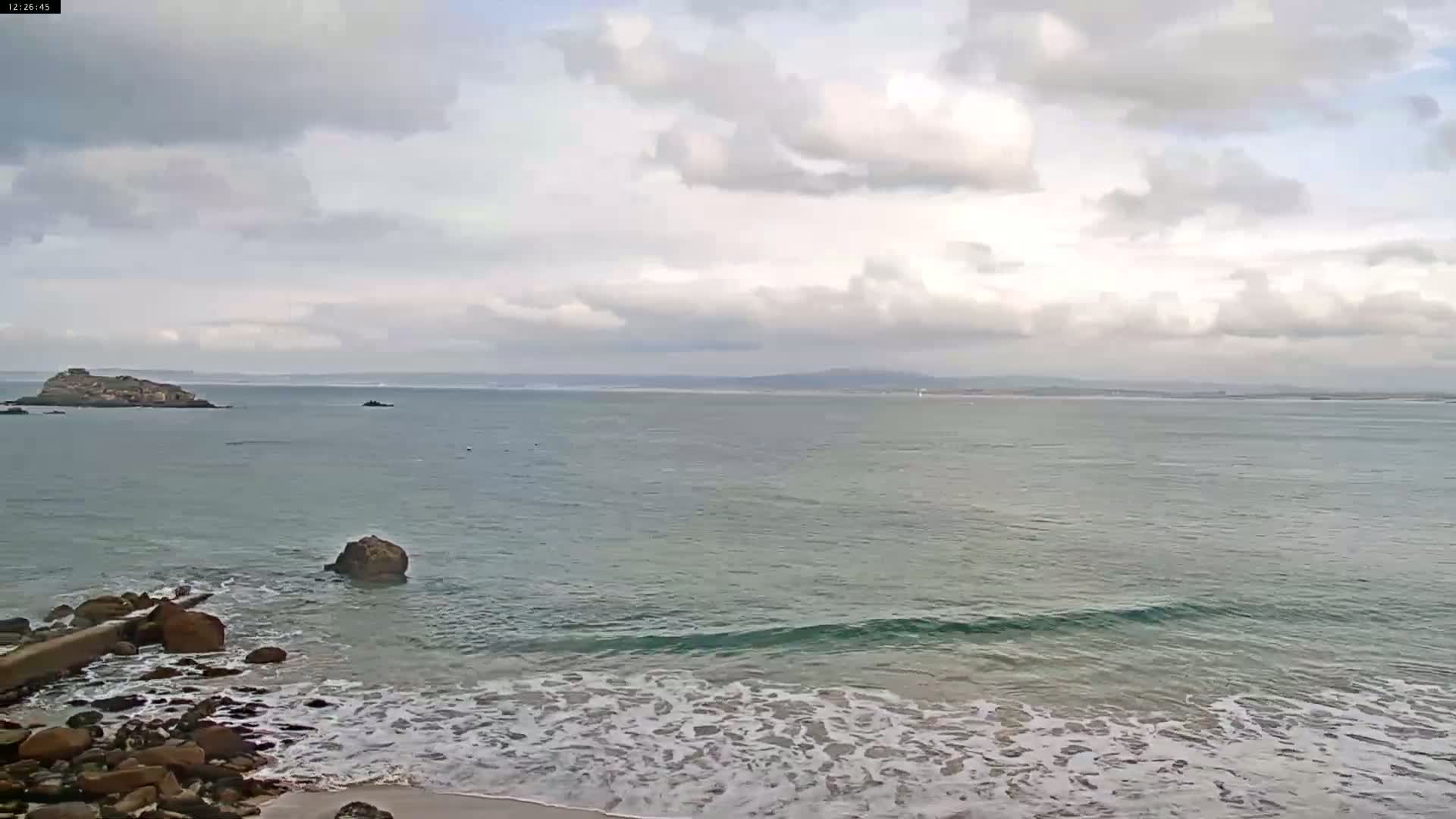 A rocky shoreline with small waves meets a calm ocean under a cloudy sky, with a distant island visible on the horizon.