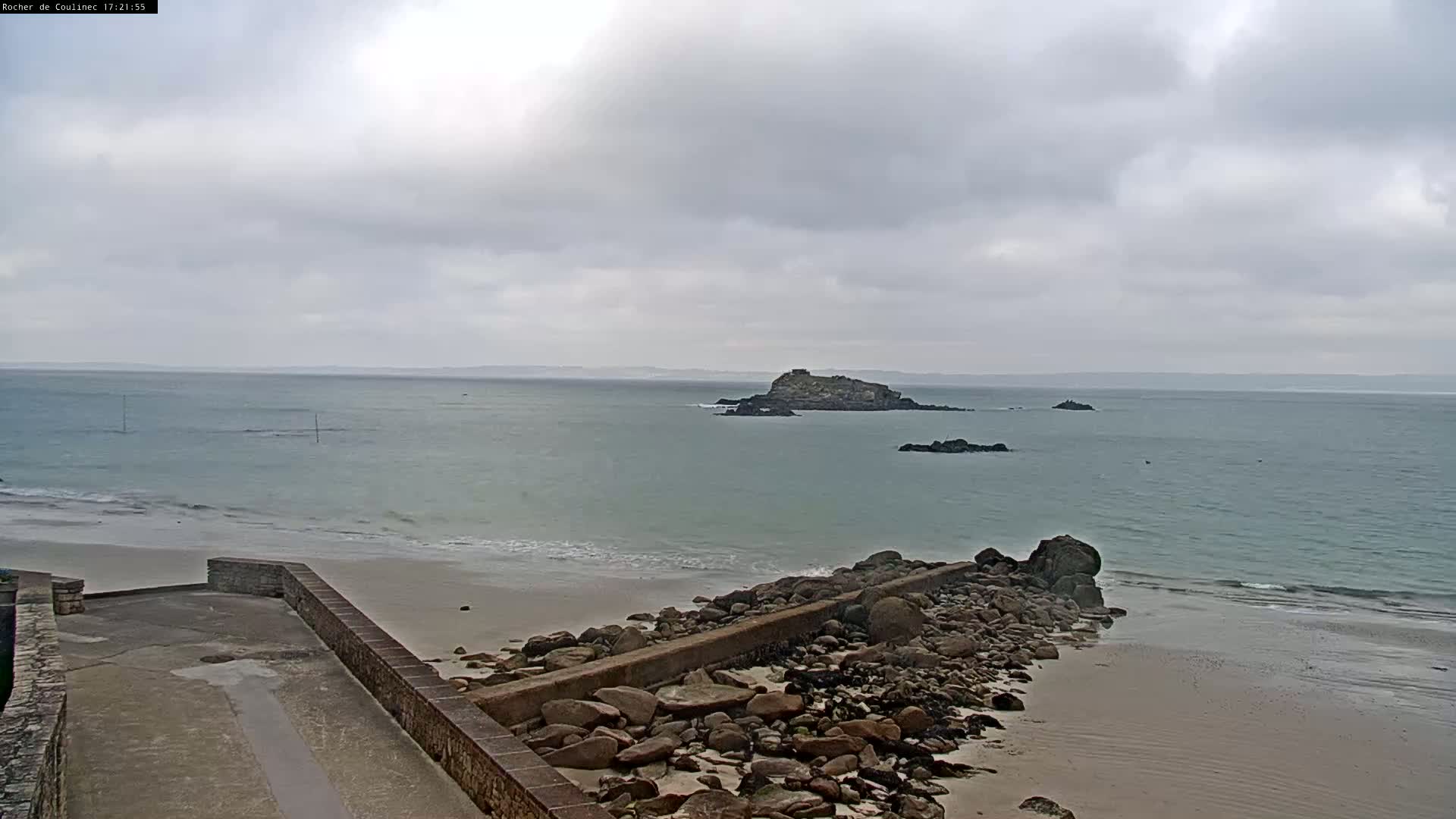 A rocky shoreline meets a calm, light-green ocean under an overcast sky, with a small island visible in the distance.