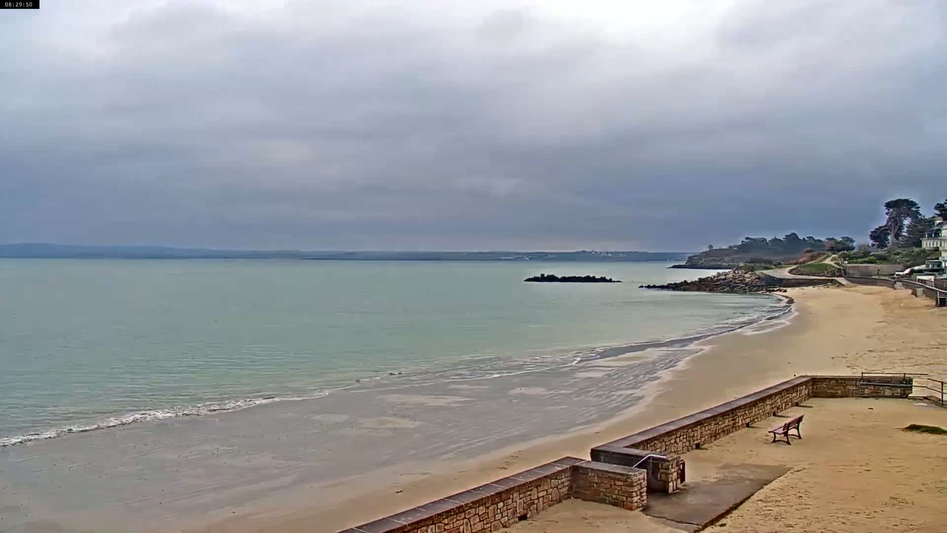 A sandy beach with calm, light-green water stretches out under a cloudy sky, bordered by a low stone wall and some vegetation.