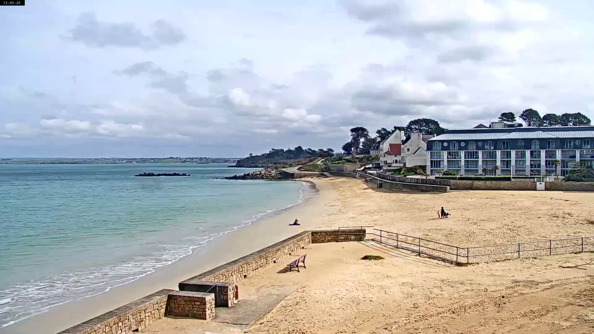 A sandy beach with calm, light-blue water laps gently at the shore, under a partly cloudy sky, next to a multi-story hotel and other buildings.