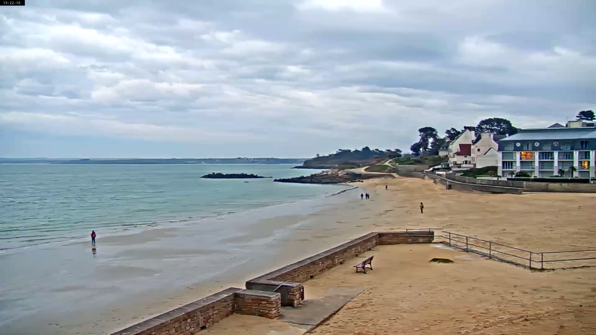 A sandy beach stretches along a calm, light-green sea under a cloudy sky, with a few people walking on the sand and buildings visible in the distance.