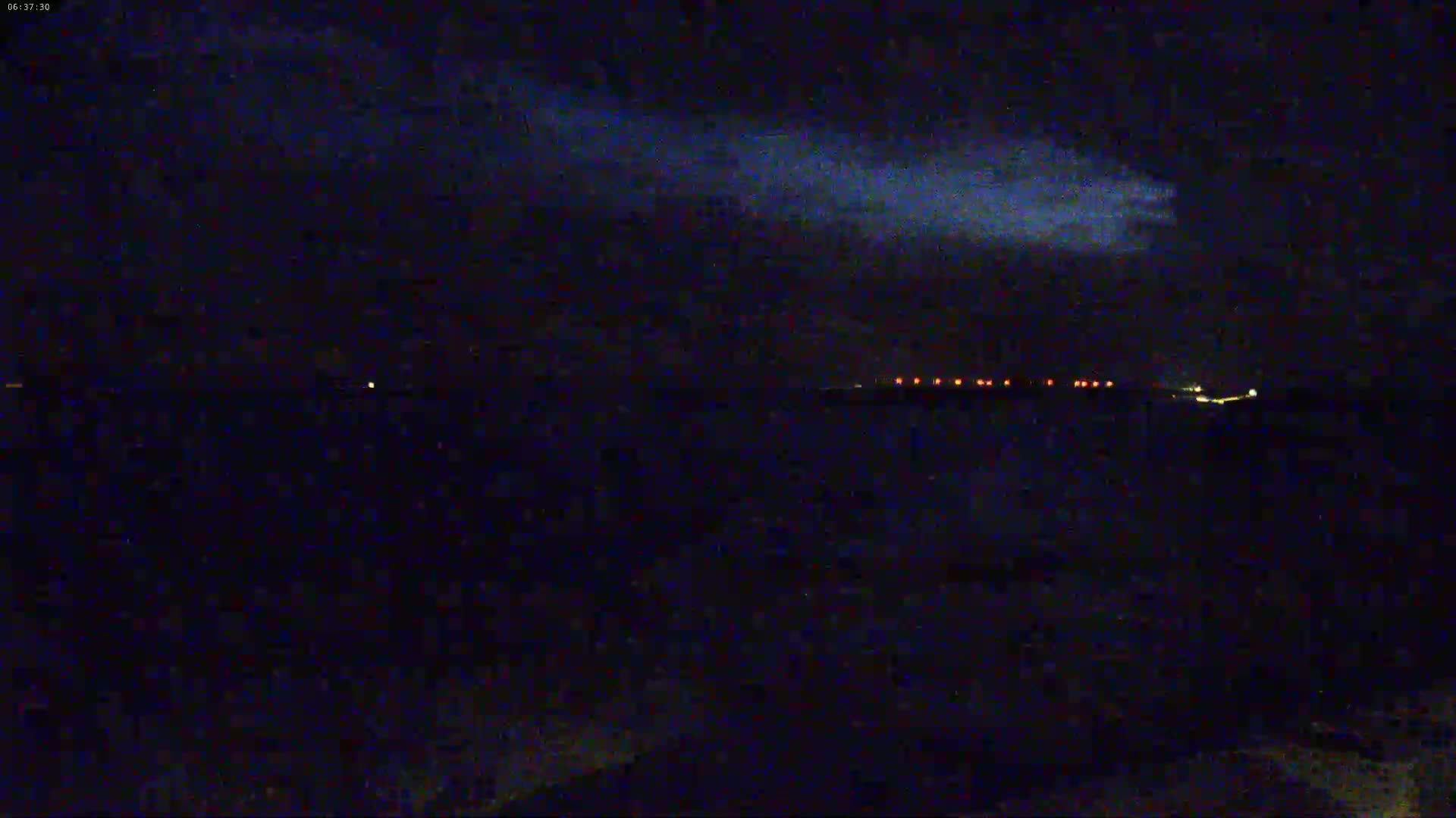 A nighttime, dark scene shows a distant line of faint, red lights under a mostly cloudy sky.