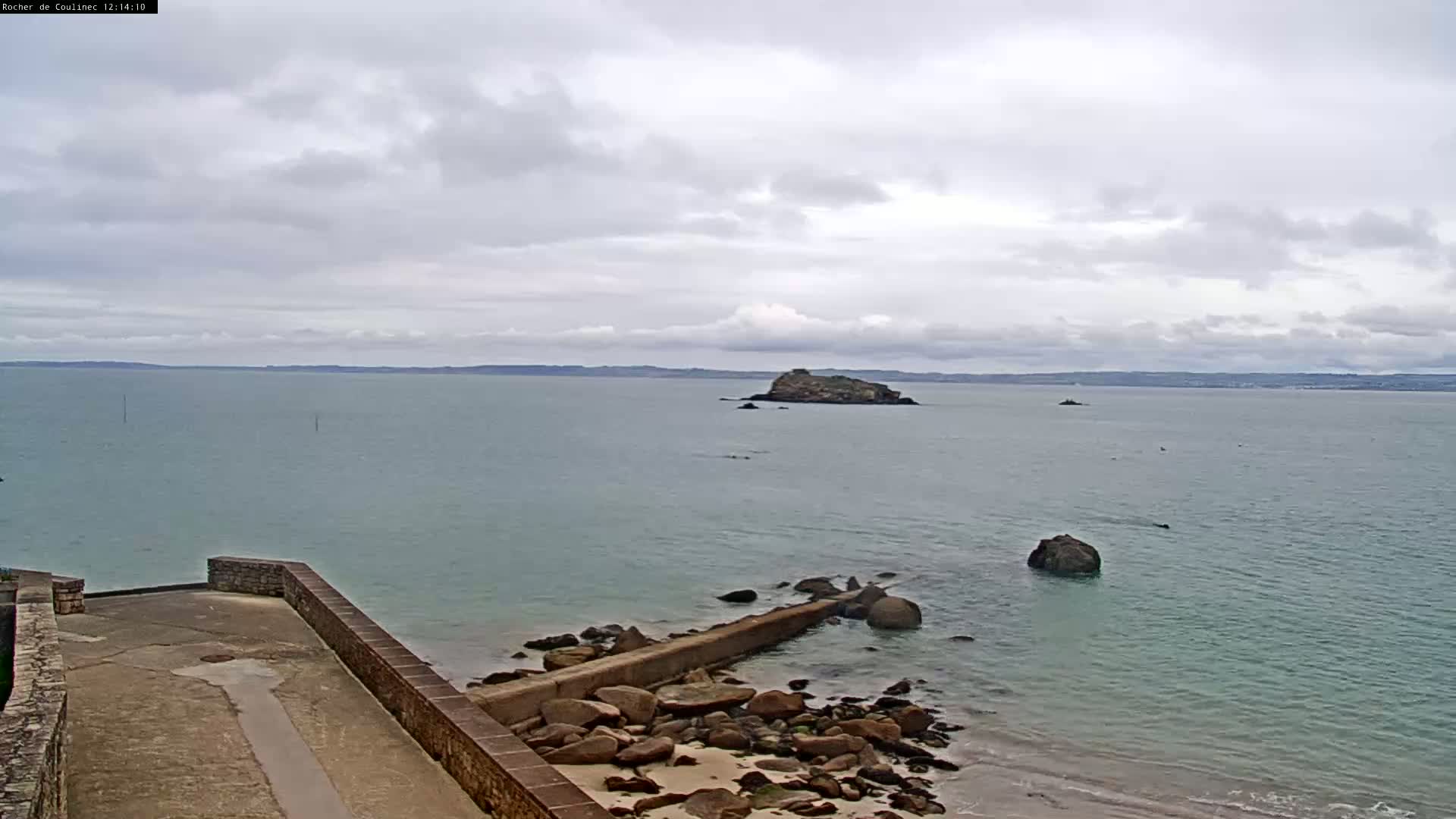 A rocky coastline with a low stone wall extends into a calm, gray sea where a small island is visible under a cloudy sky.
