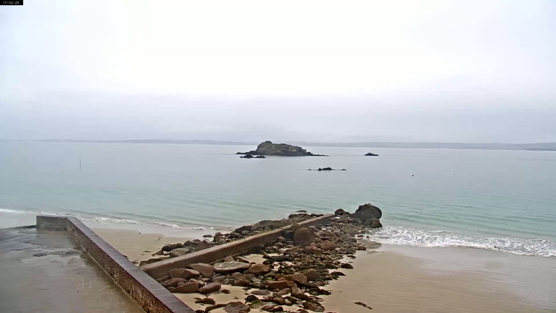 A rocky outcropping sits in a calm, light-green ocean under an overcast sky, near a sandy beach with a low stone wall.