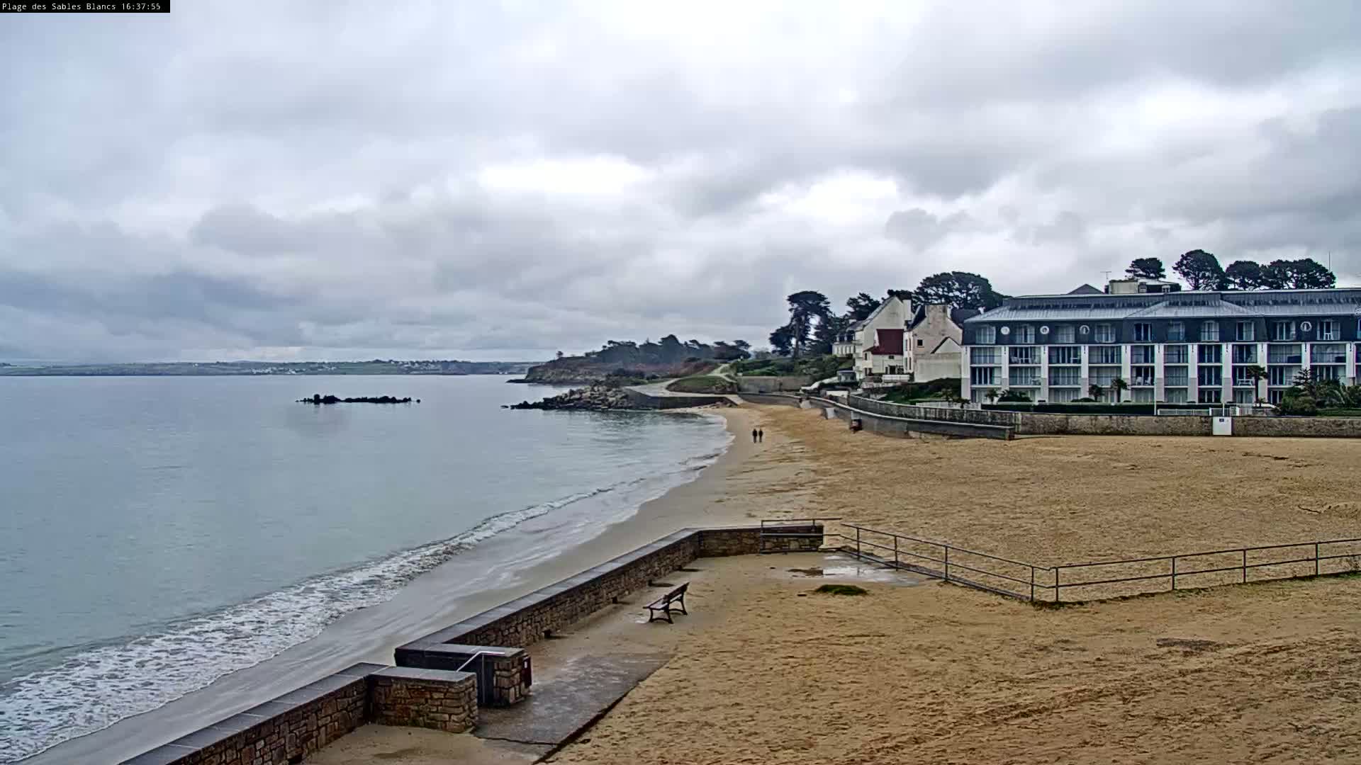 A sandy beach with a low stone wall, gentle waves, and a multi-story building overlooks calm water under an overcast sky.