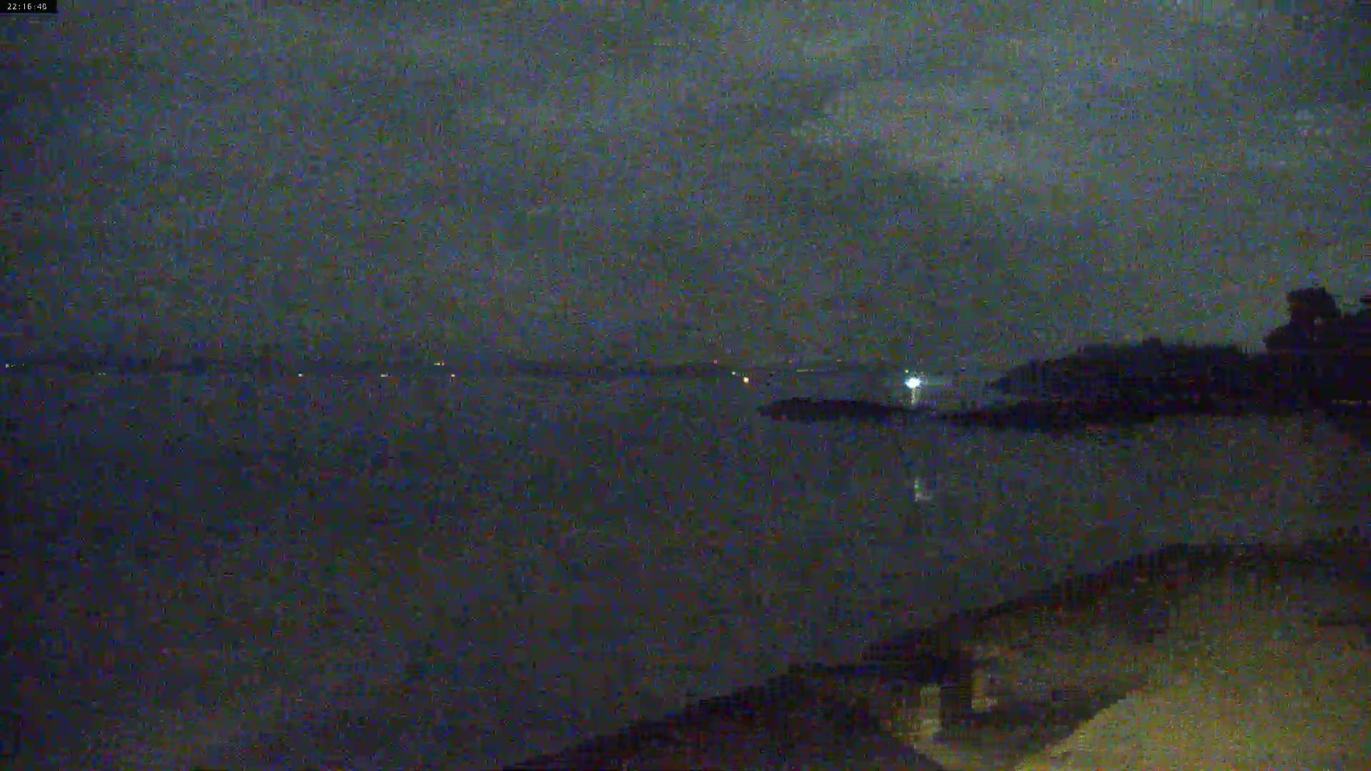 A dark, nighttime view of a calm body of water under a cloudy sky shows distant lights across the water and a shoreline with dark trees in the foreground.