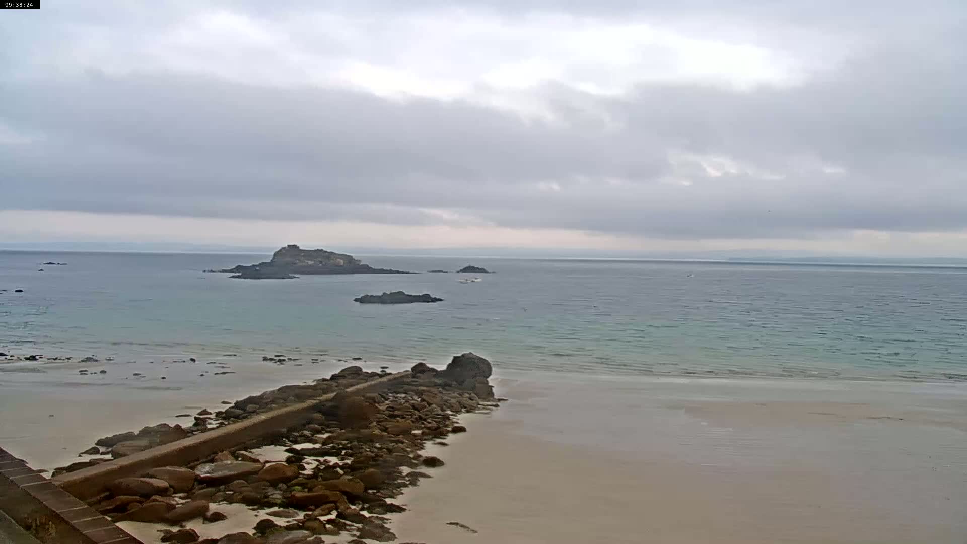 A rocky shoreline with a light-colored sandy beach meets a calm, light-teal ocean under an overcast sky.