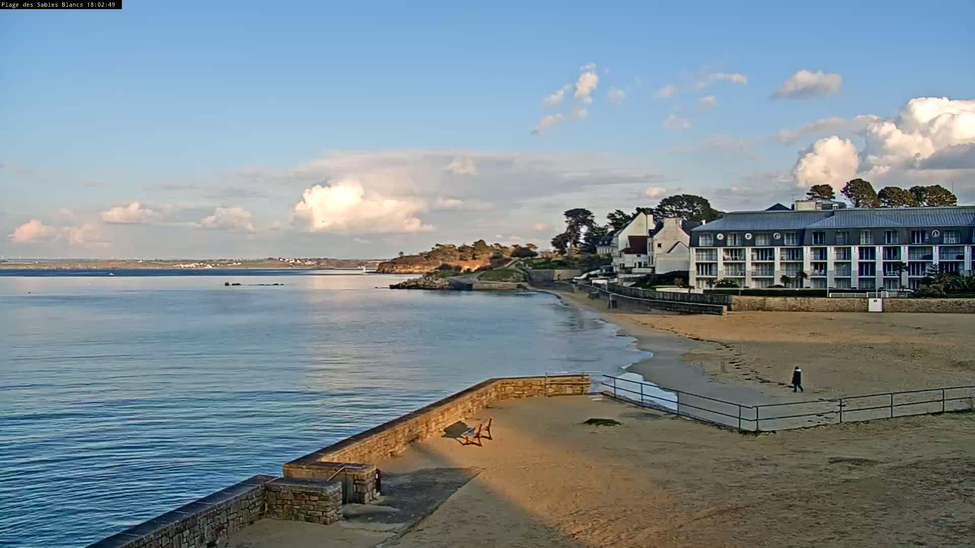 A sandy beach with a low stone wall and a large hotel overlooks a calm body of water under a partly cloudy sky.