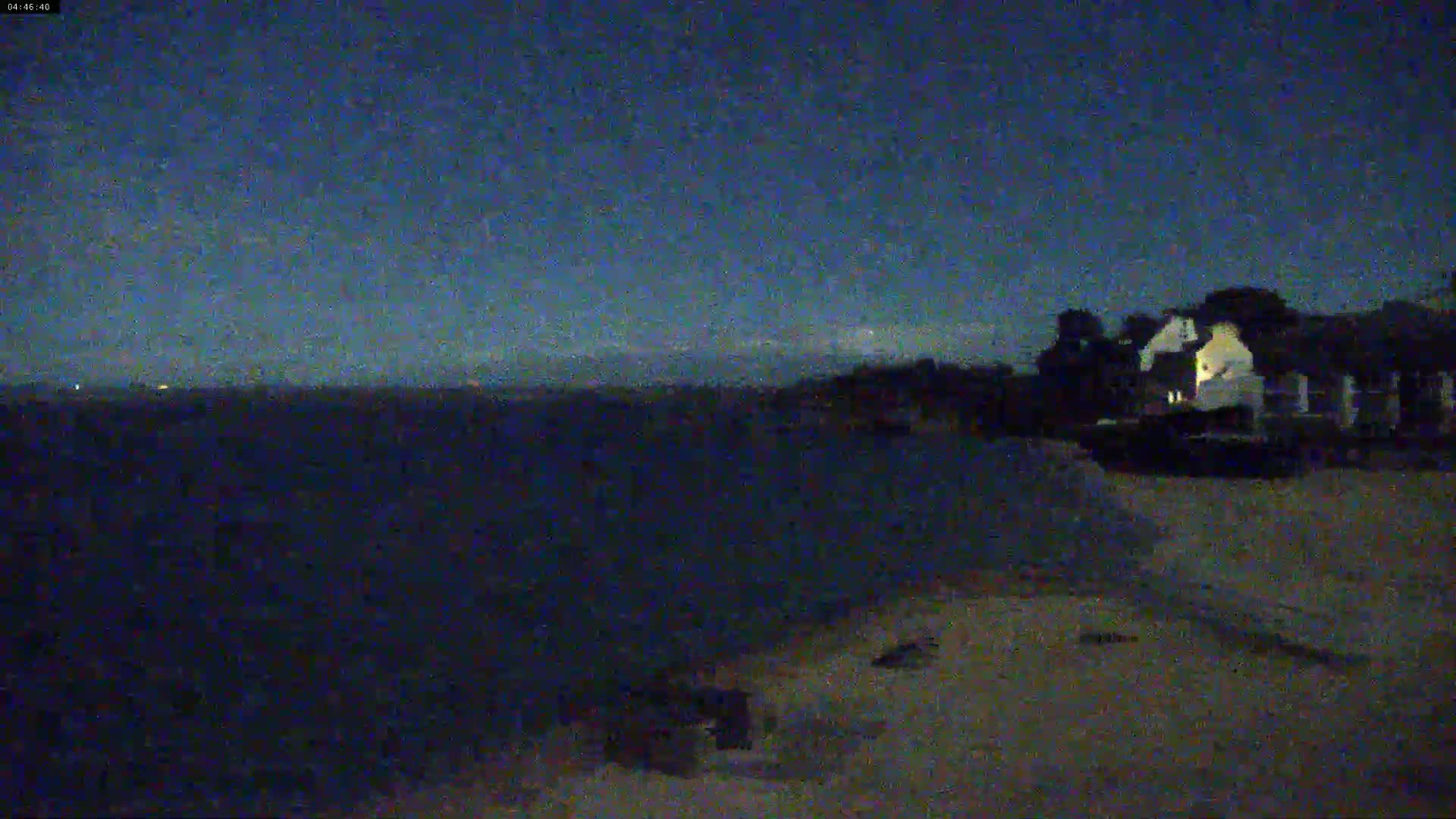 A dark, nighttime scene shows a body of water and a building near a shoreline under a clear, dark blue sky.