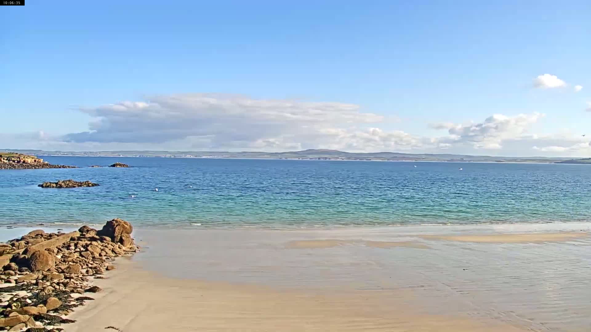 A sandy beach slopes gently into calm, teal-colored water under a mostly sunny sky with scattered clouds.