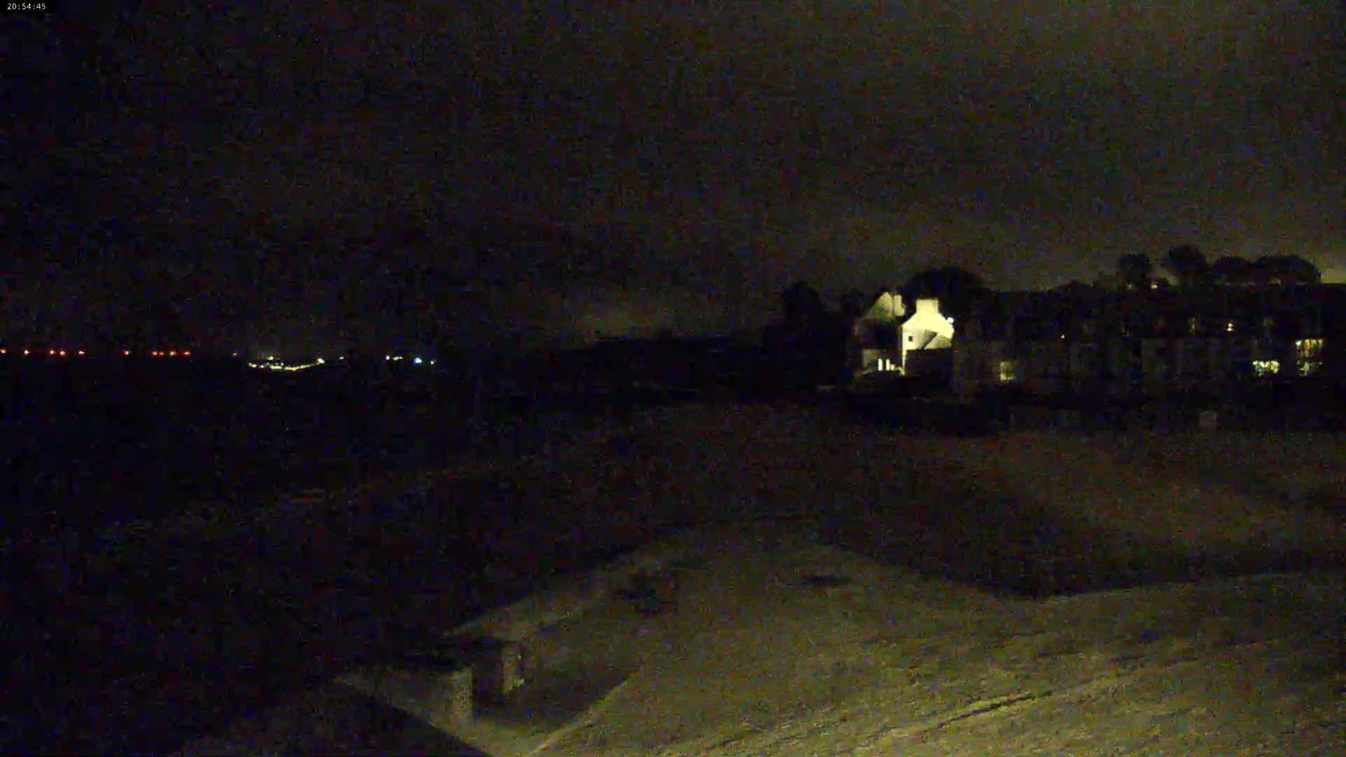 A nighttime view shows a snow-covered ground in the foreground with dimly lit buildings and distant lights visible under a dark, cloudy sky.