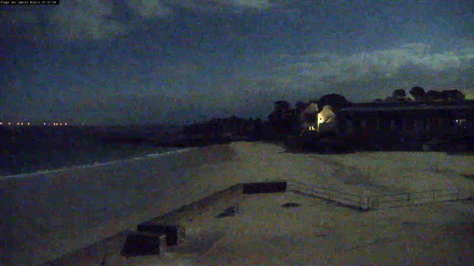 A dark, sandy beach at night is partially illuminated by lights from a building beyond a low wall, under a mostly cloudy sky.