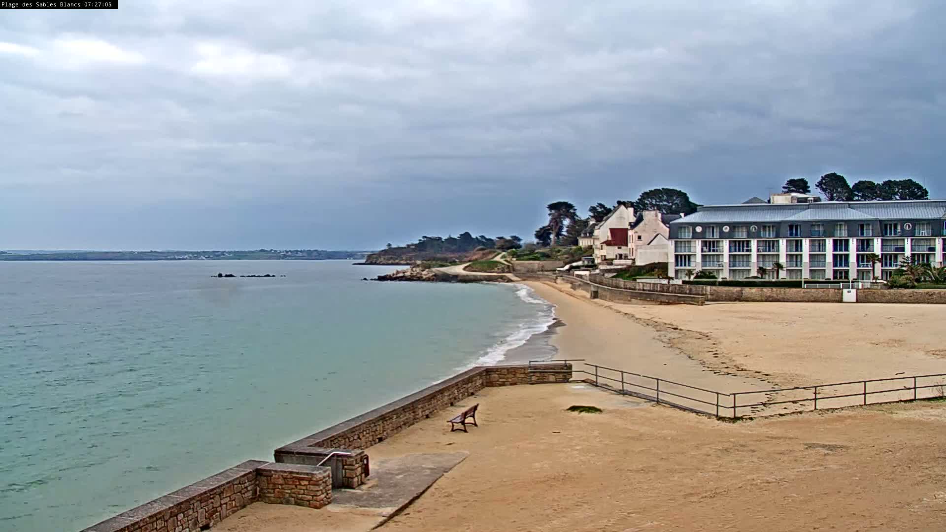 A sandy beach with a low stone wall and a multi-story building is visible under an overcast sky.