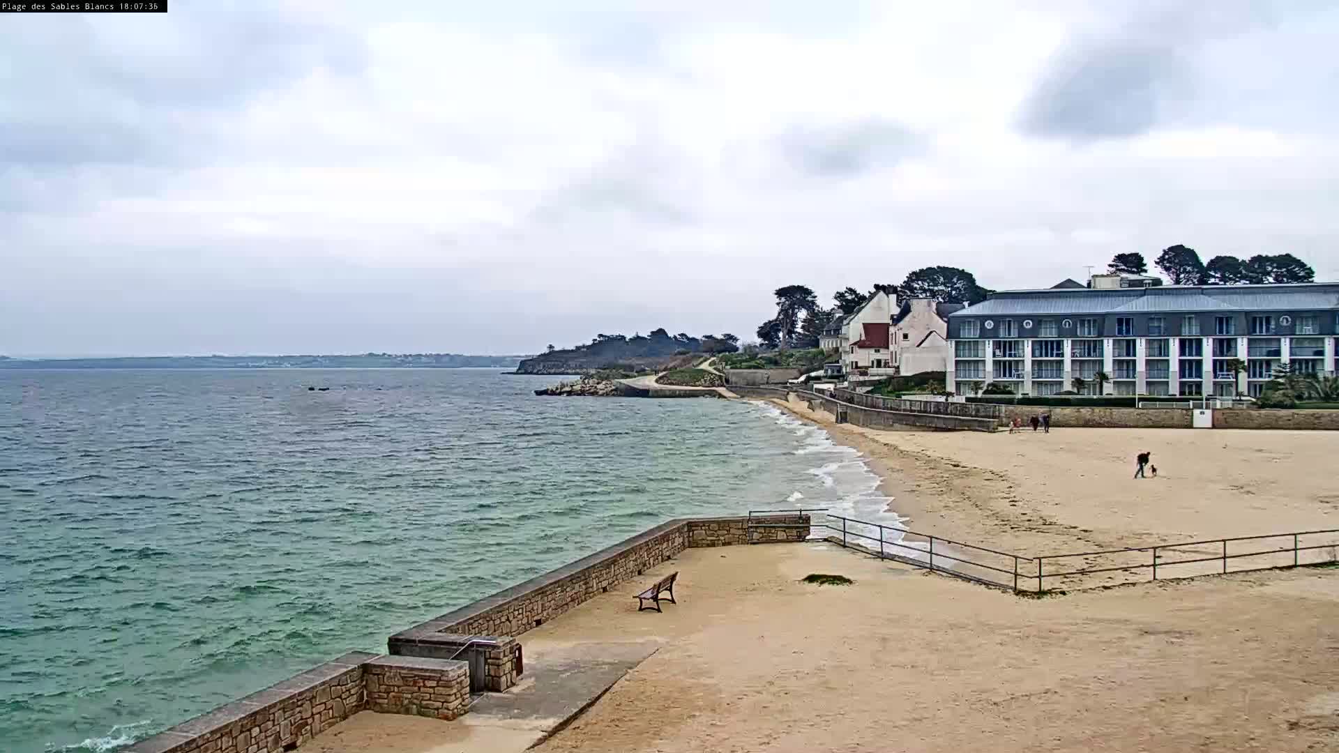 A sandy beach with a stone wall and a large building overlooks a calm, greenish-blue body of water under a cloudy sky.